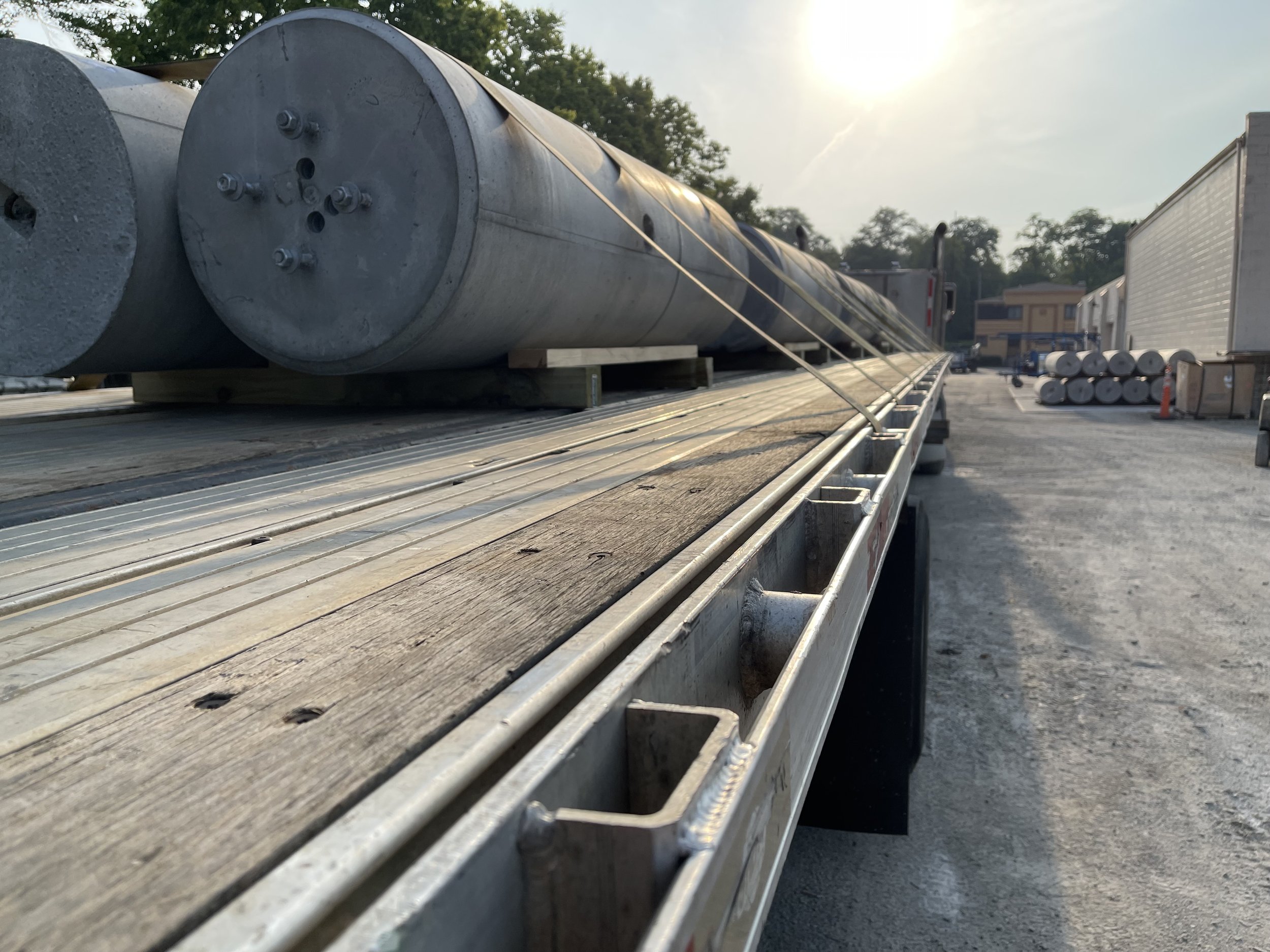 A flatbed truck carrying large cylindrical metal pipes in an industrial area during sunset.