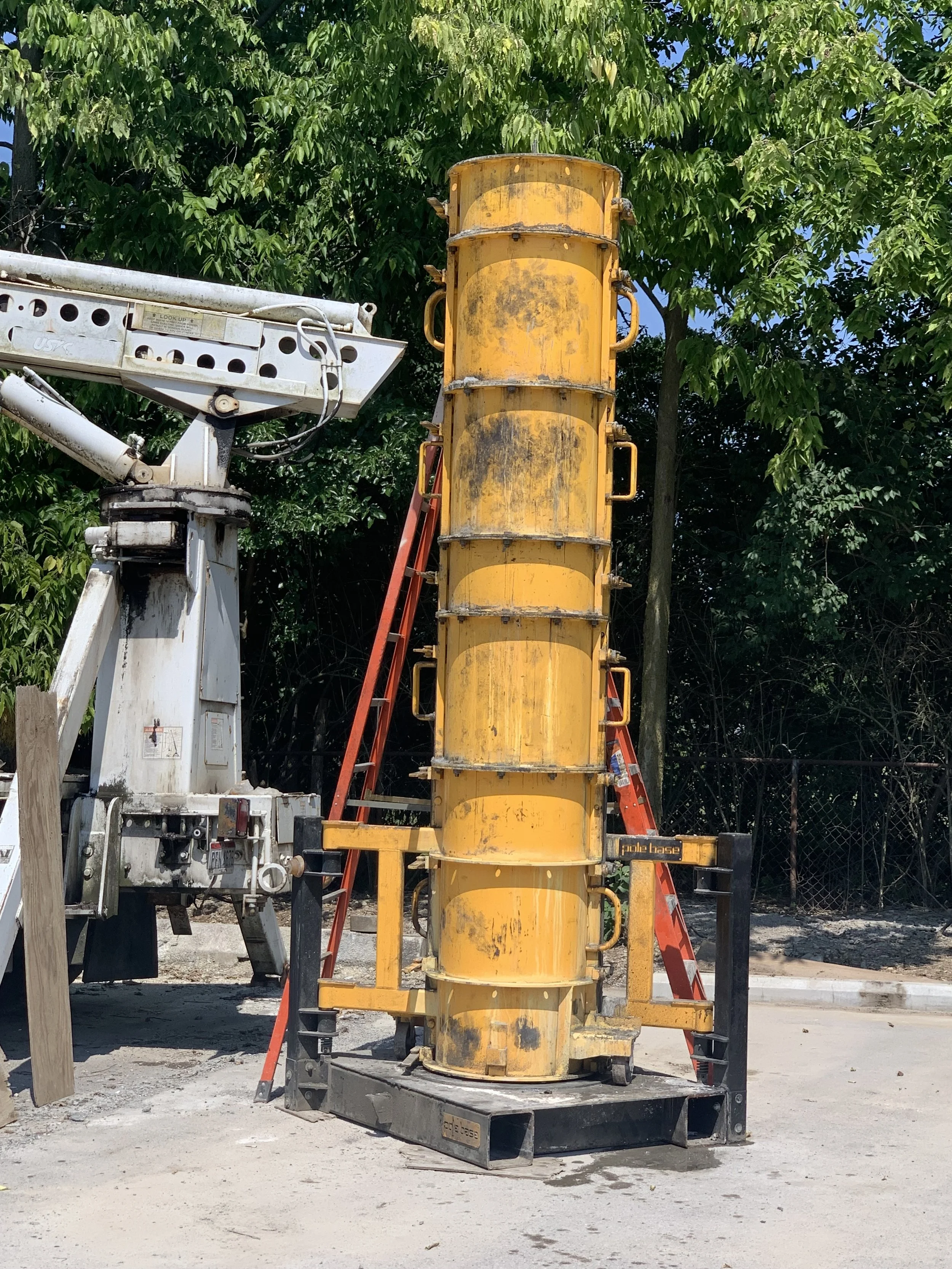 A large yellow construction safety barrier with multiple handles, placed on a black stand, with a red ladder leaning against it, outdoor setting with green trees in background.