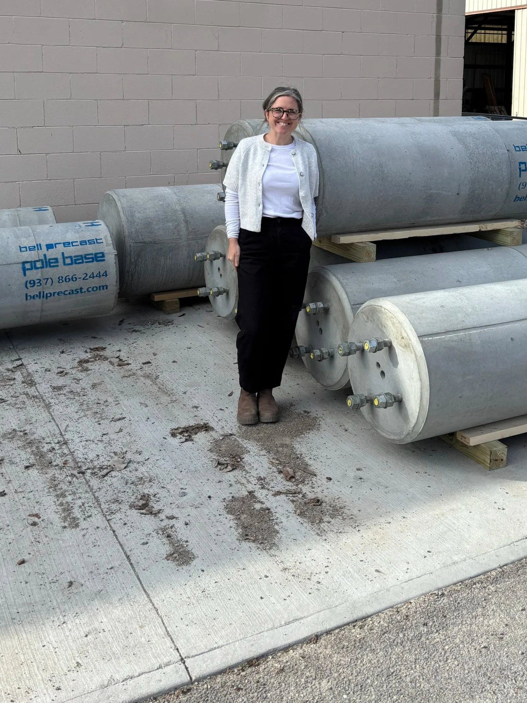 A woman standing outdoors next to large metal tanks labeled 'pole base' with a smile, in front of a beige brick wall and a warehouse building under a blue sky with some clouds.