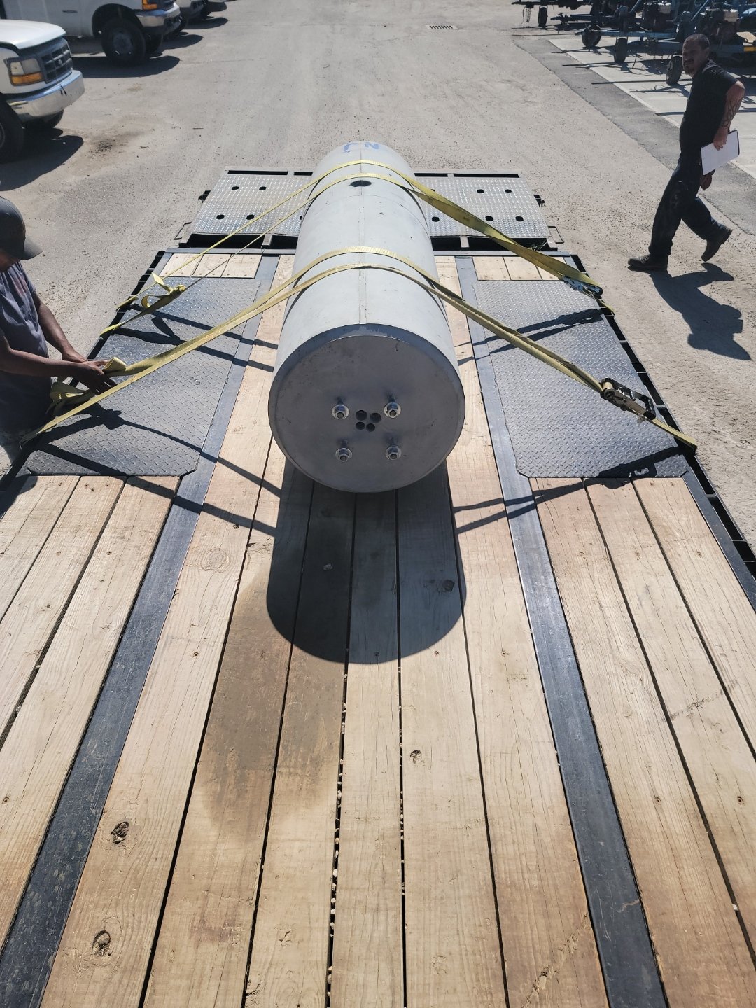A large cylindrical metal object secured on a flatbed truck with yellow straps in a parking lot.