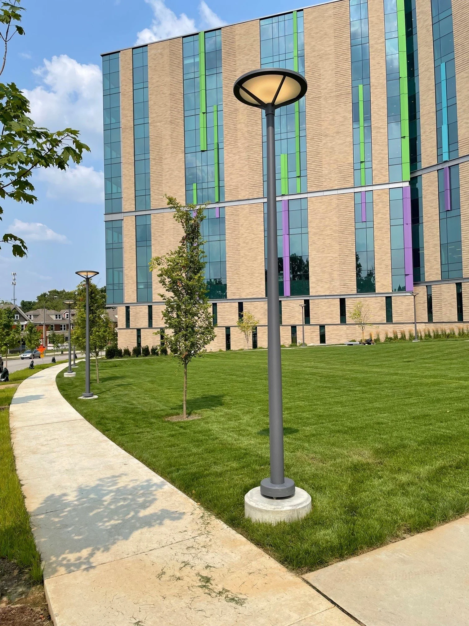 Modern multi-story building with colorful vertical accents and reflective glass windows, adjacent to a landscaped lawn with young trees, concrete sidewalks, and curved pathway, under partly cloudy sky.