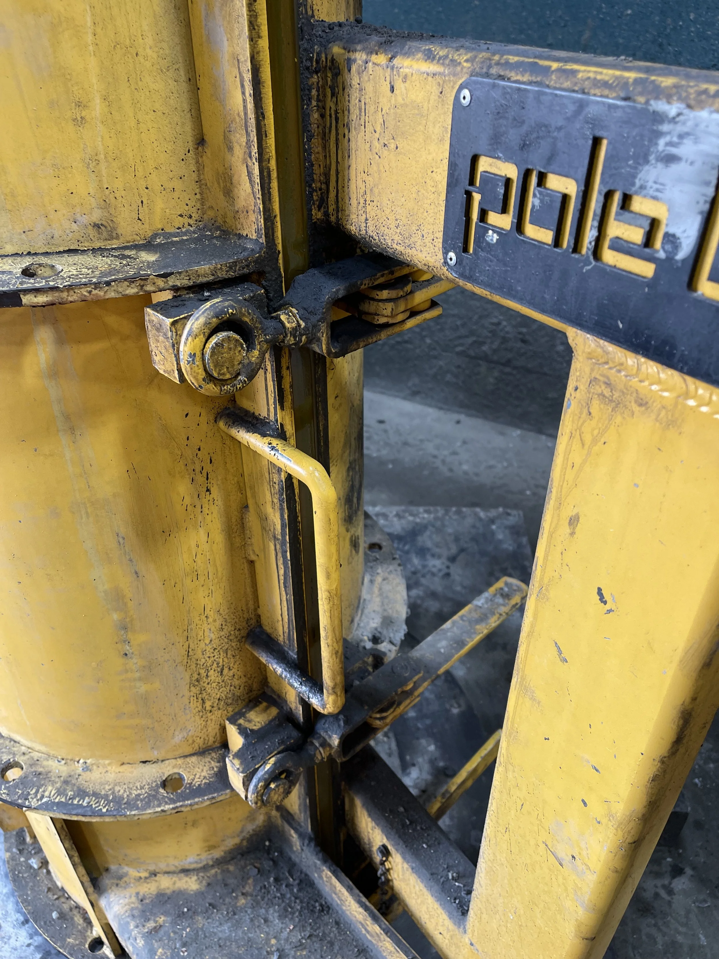 Close-up of a yellow industrial machine with a black metal label with the word 'poles' on it, showing metal parts, hinges, and mounting hardware.