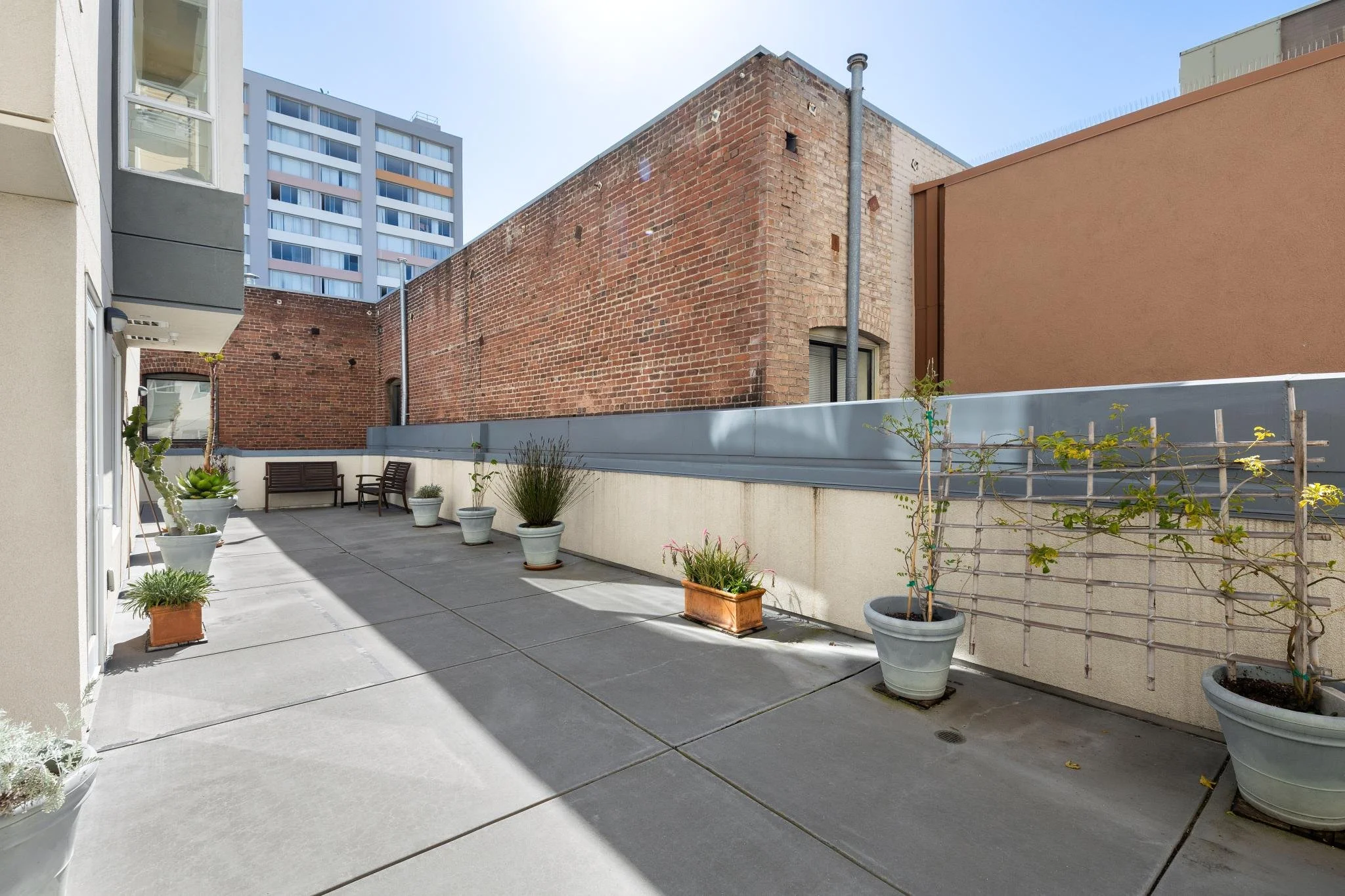 View of an outdoor balcony with potted plants, a wooden bench, and two chairs, with brick and modern buildings in the background on a sunny day.