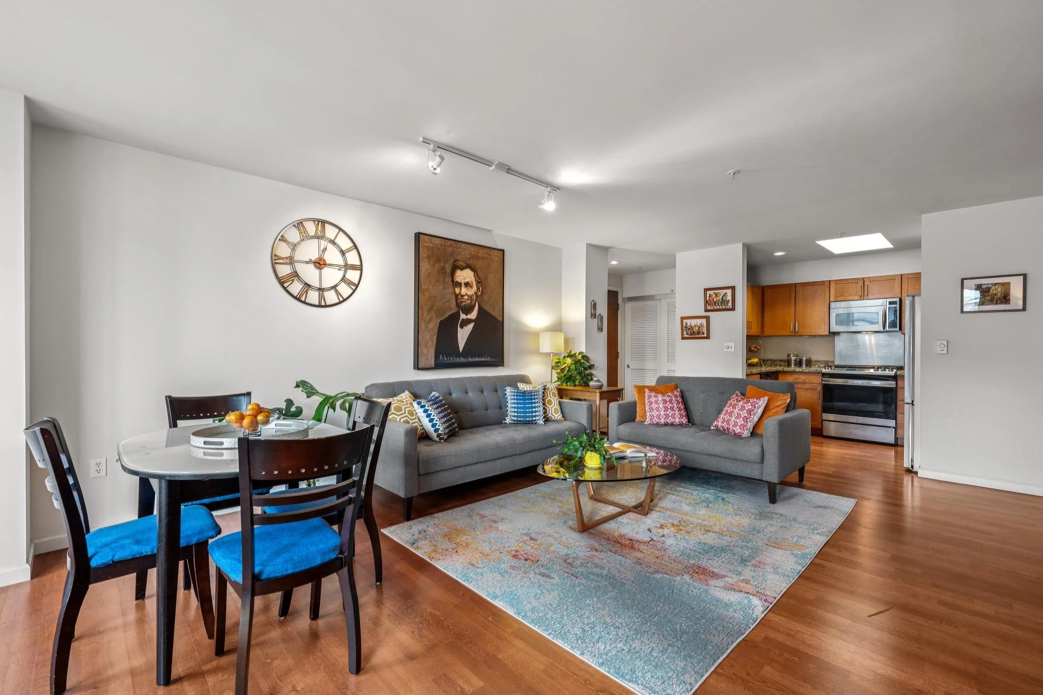 Open-concept living room and dining area with hardwood floors, two gray sofas with colorful pillows, a round glass coffee table, a wooden dining table with four chairs, a large wall clock, a portrait, and the kitchen in the background.