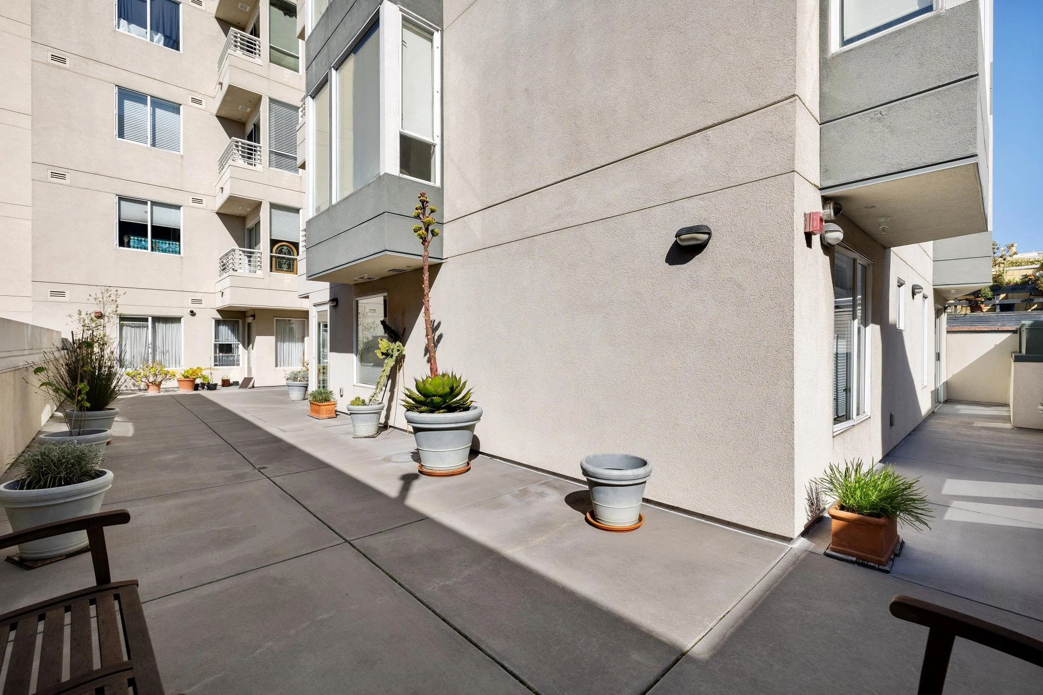 Exterior view of a modern apartment building's outdoor patio area with potted plants, a bench, and windows with balconies.