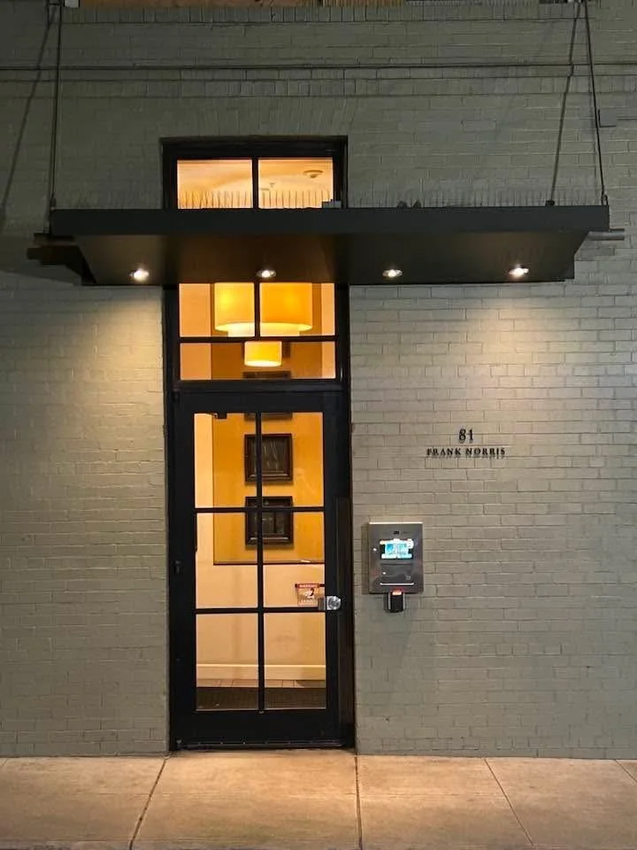 Exterior view of a modern apartment building entrance with a black framed glass door, a wall-mounted mail and package locker, and the address 81 Frank Norris displayed on the wall. The building has a brick facade and a small overhang with built-in li
