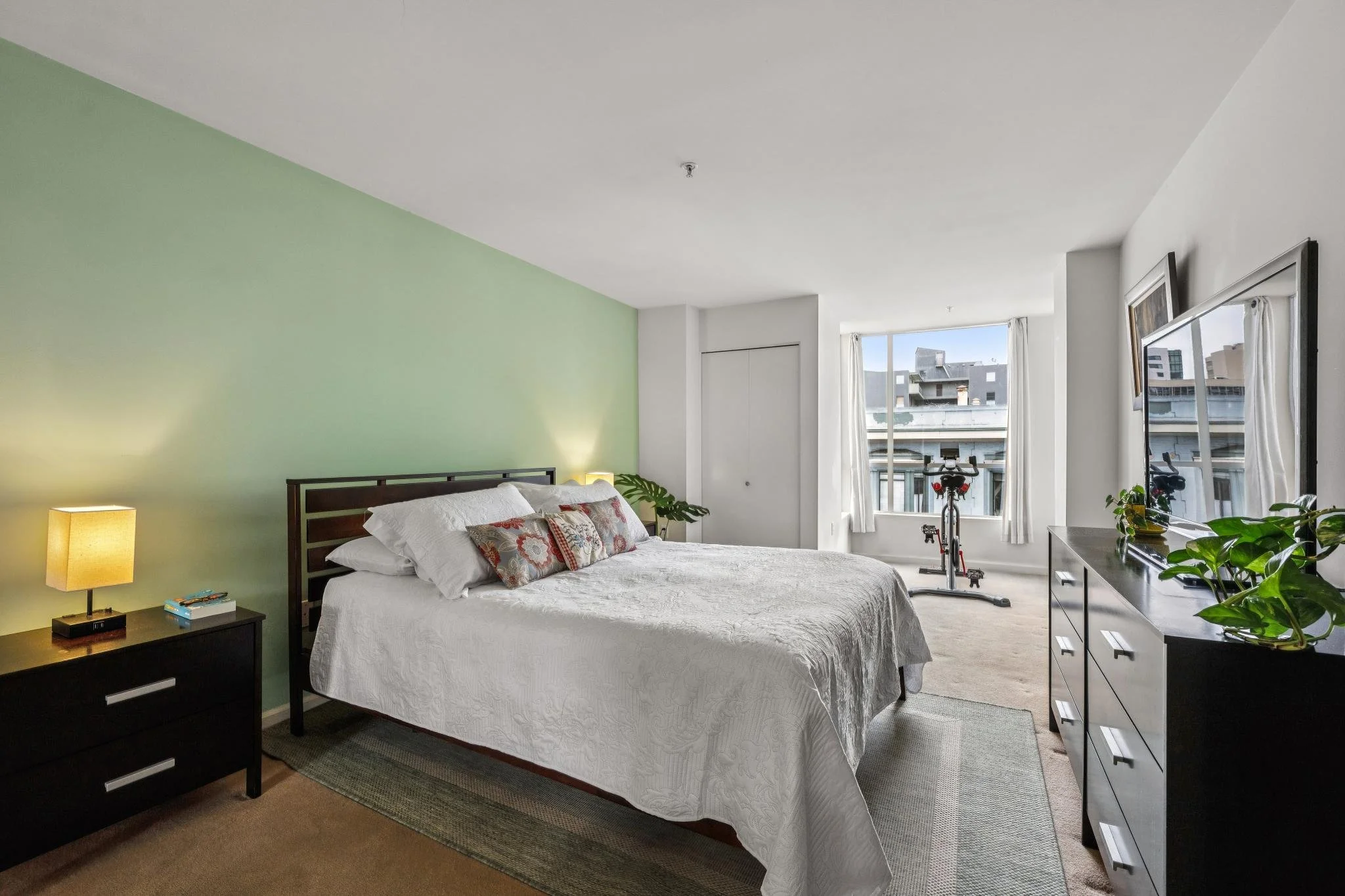 A bedroom with a green accent wall, a white bed with pillows, two nightstands with lamps, a dresser with plants, and a window with curtains overlooking city buildings. There's also a stationary exercise bike near the window.