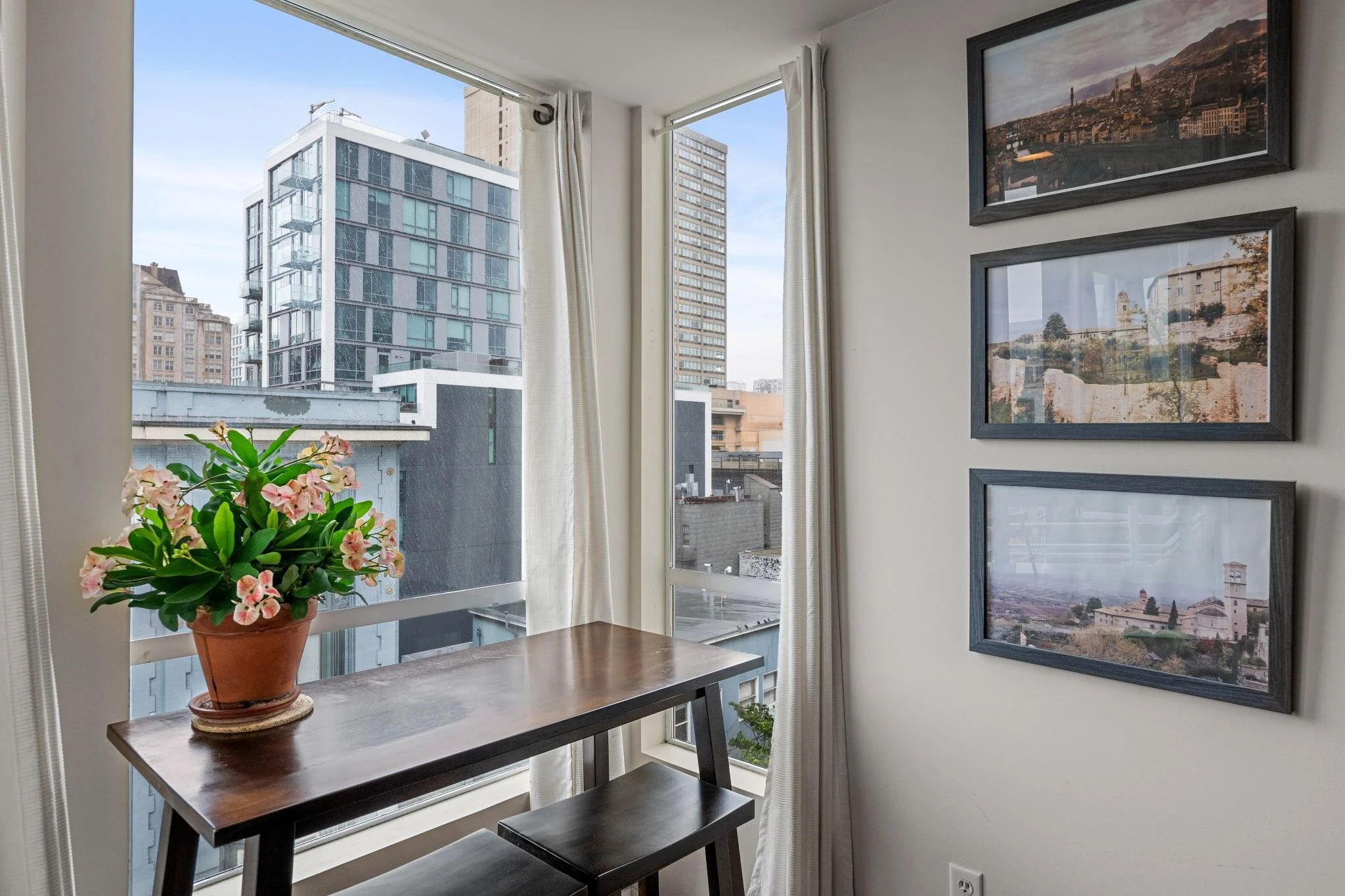 City view from an apartment window with a wooden table and stool, a potted plant, and framed pictures on the wall.