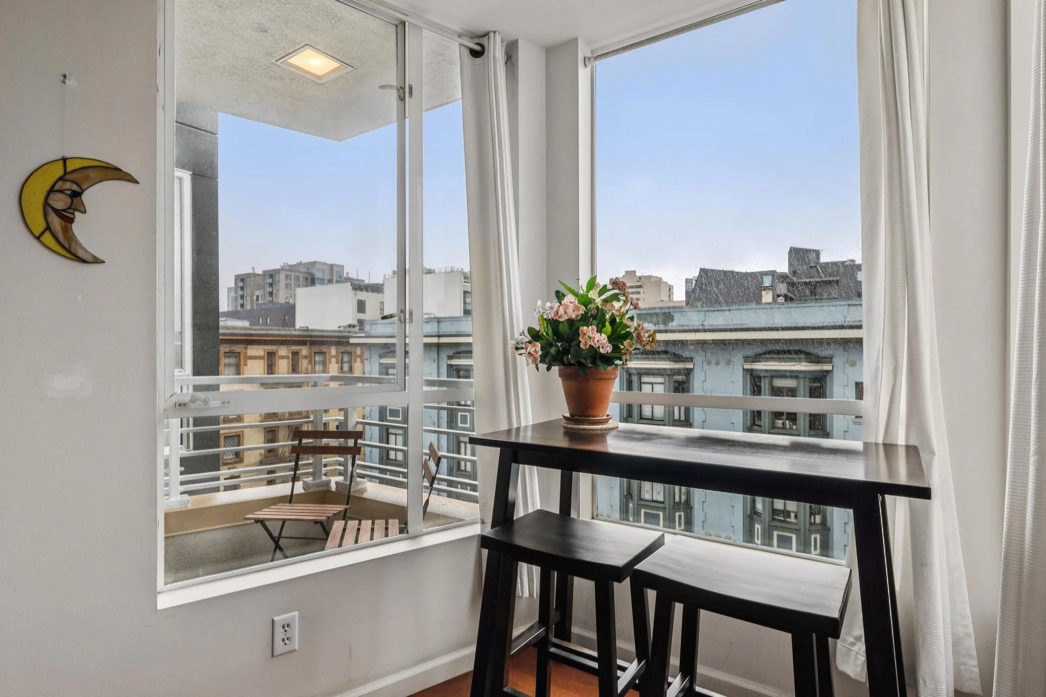 A small balcony with a wooden table and two stools, a flower pot with pink flowers, and large windows with white curtains showing city buildings outside.