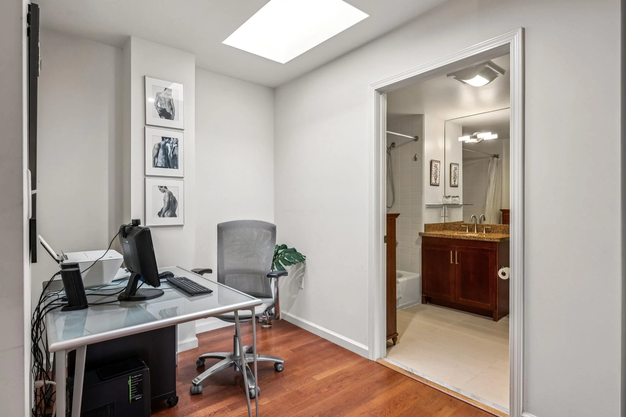 An office with a desk, computer, and office chair, located next to a bathroom with a vanity, mirror, and shower.