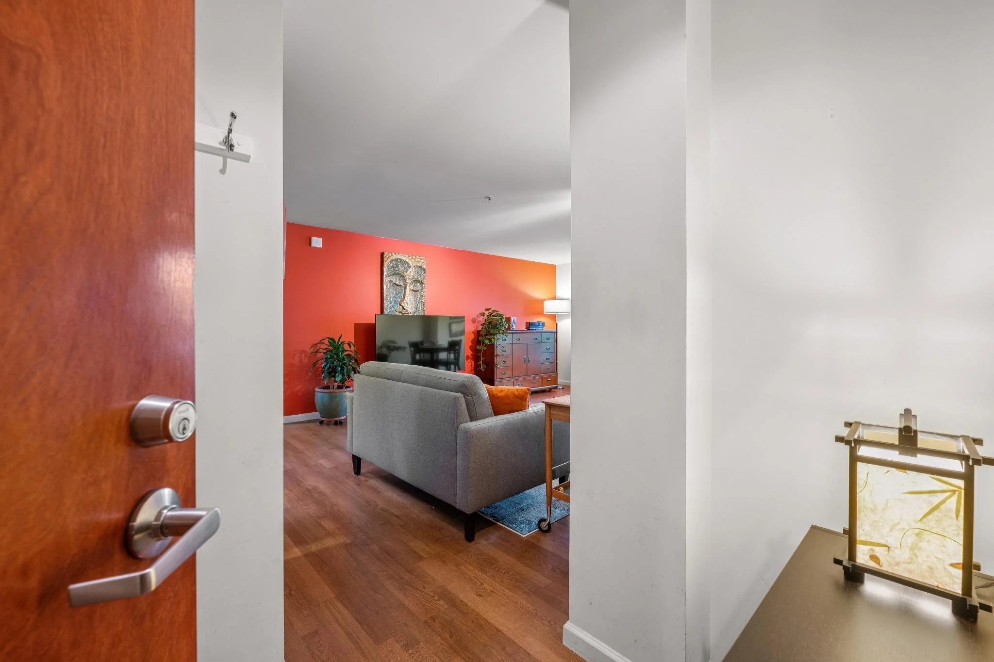 View of a living room seen through an open doorway, featuring a red accent wall, a tv, a gray sofa, a wooden sideboard, and indoor plants.