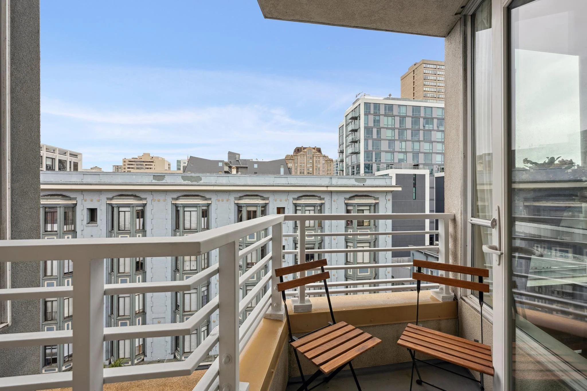 Small balcony with two wooden chairs and white railing, overlooking city buildings and blue sky.