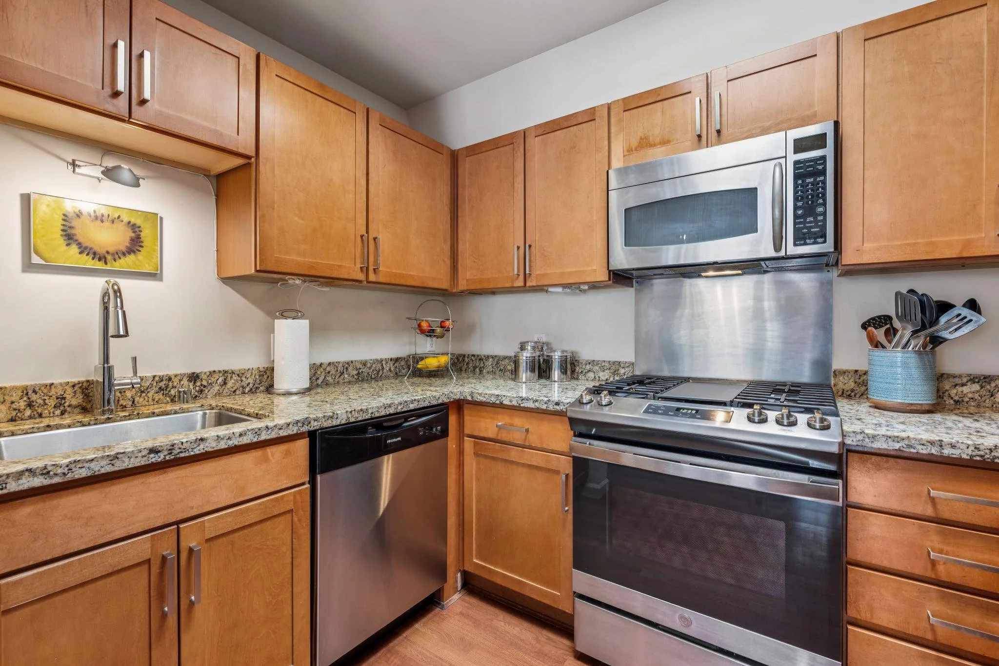 Kitchen with wooden cabinets, granite countertops, stainless steel appliances including a microwave, oven, dishwasher, and a sink with a modern faucet. There are some small containers on the counter, a paper towel roll, and a framed fruit-themed artw