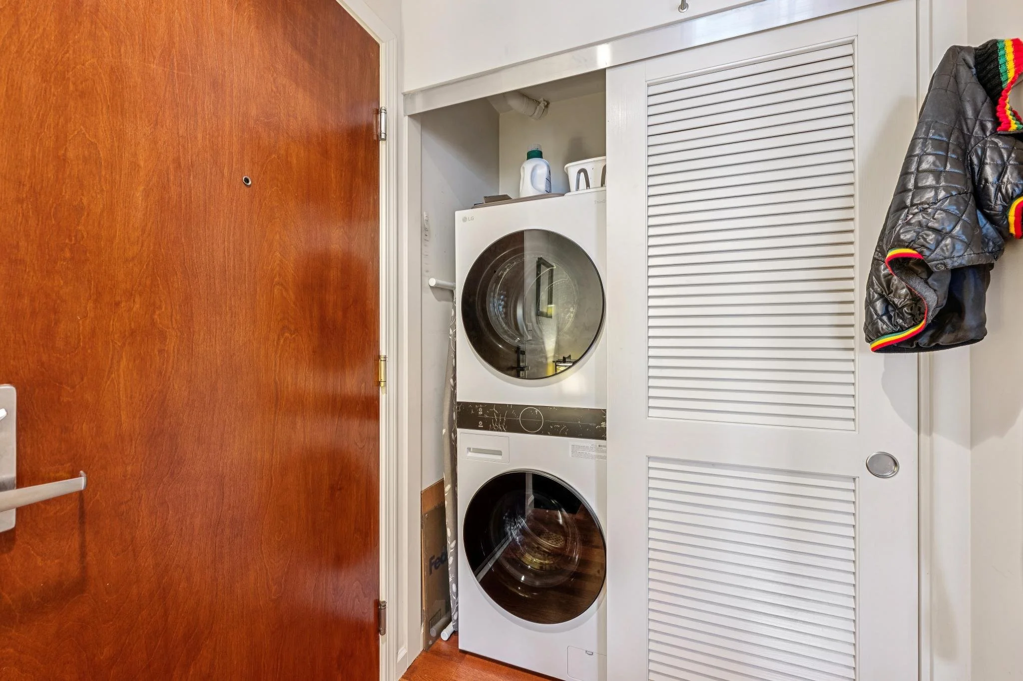 Stacked washer and dryer unit behind a louvered closet door in a laundry area with a wooden door on the left and a coat hanging on the wall to the right.