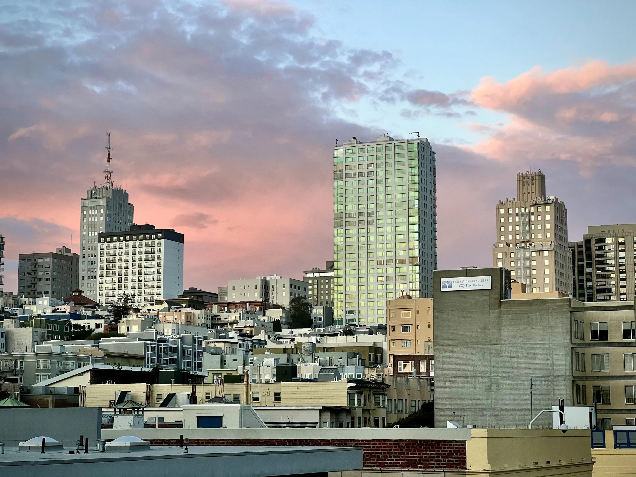 City skyline with tall buildings at sunset, colorful clouds in the sky, and various residential and commercial structures in the foreground.