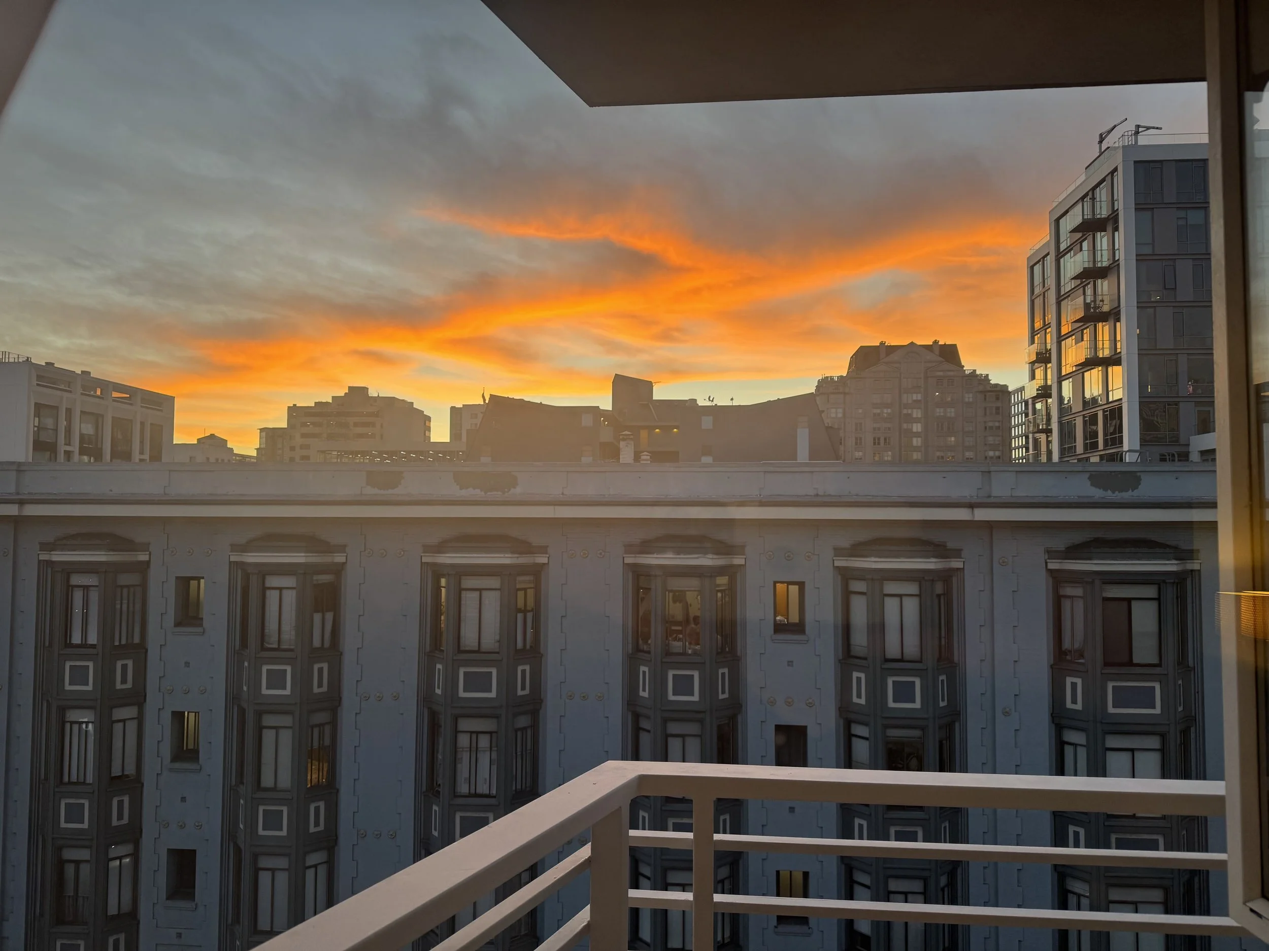 Sunset over a cityscape, viewed from a balcony. Buildings of various heights and architectural styles are visible, with the sky painted in shades of orange and yellow.