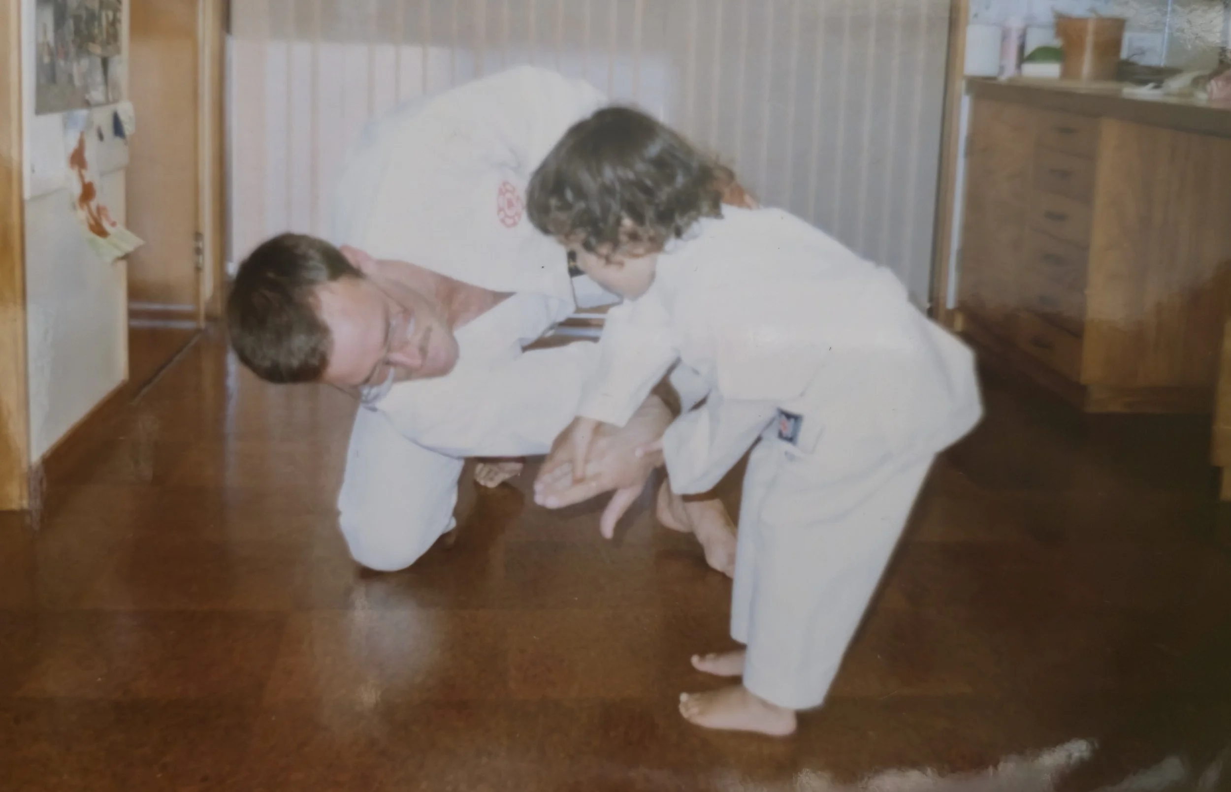 A man and a young girl practicing a martial arts move on a wooden floor in a home setting, with wooden furniture and a striped wall in the background.