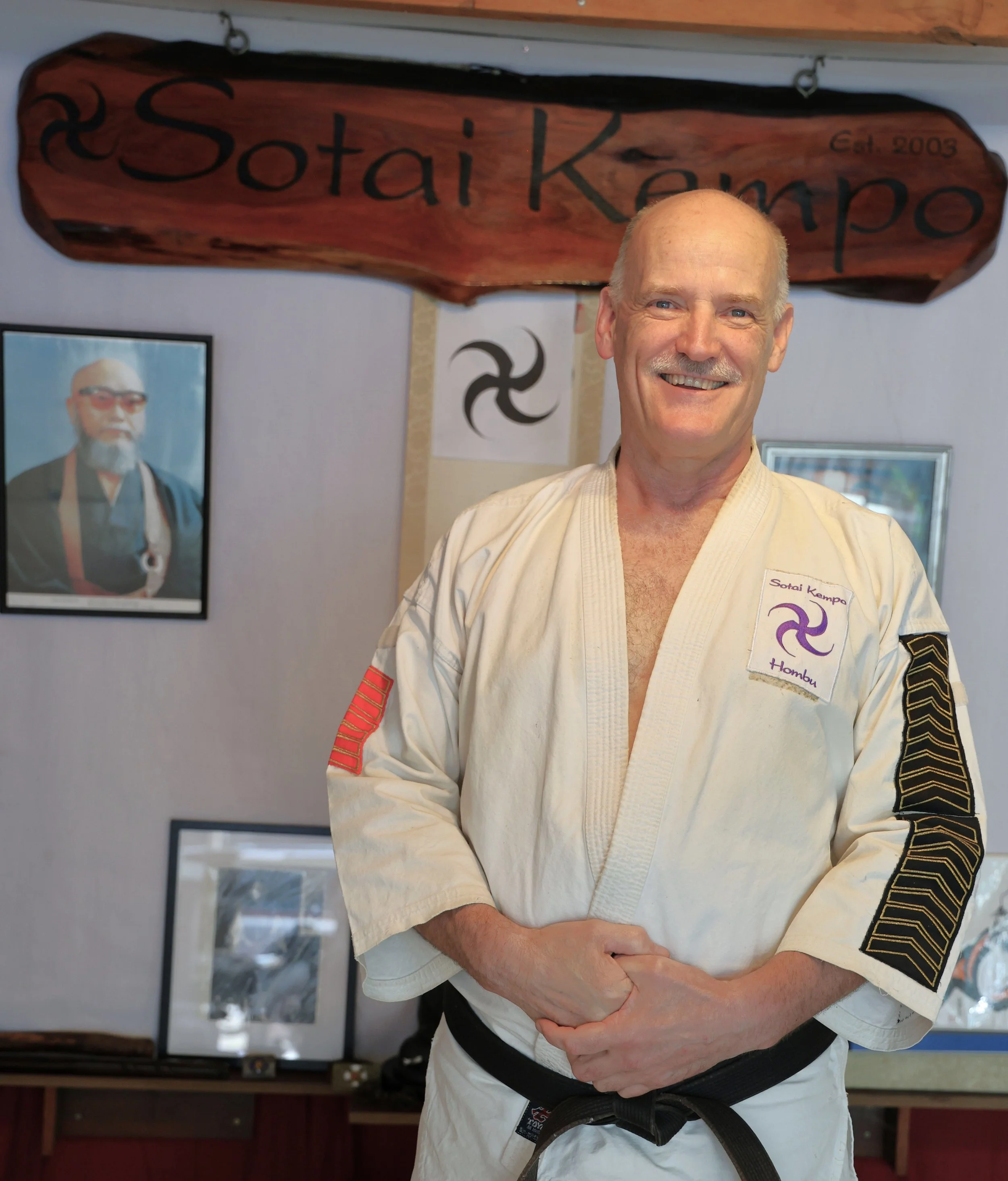 A smiling man in a white martial arts uniform with black belt, standing in front of a sign that reads "Sotai Kempo".