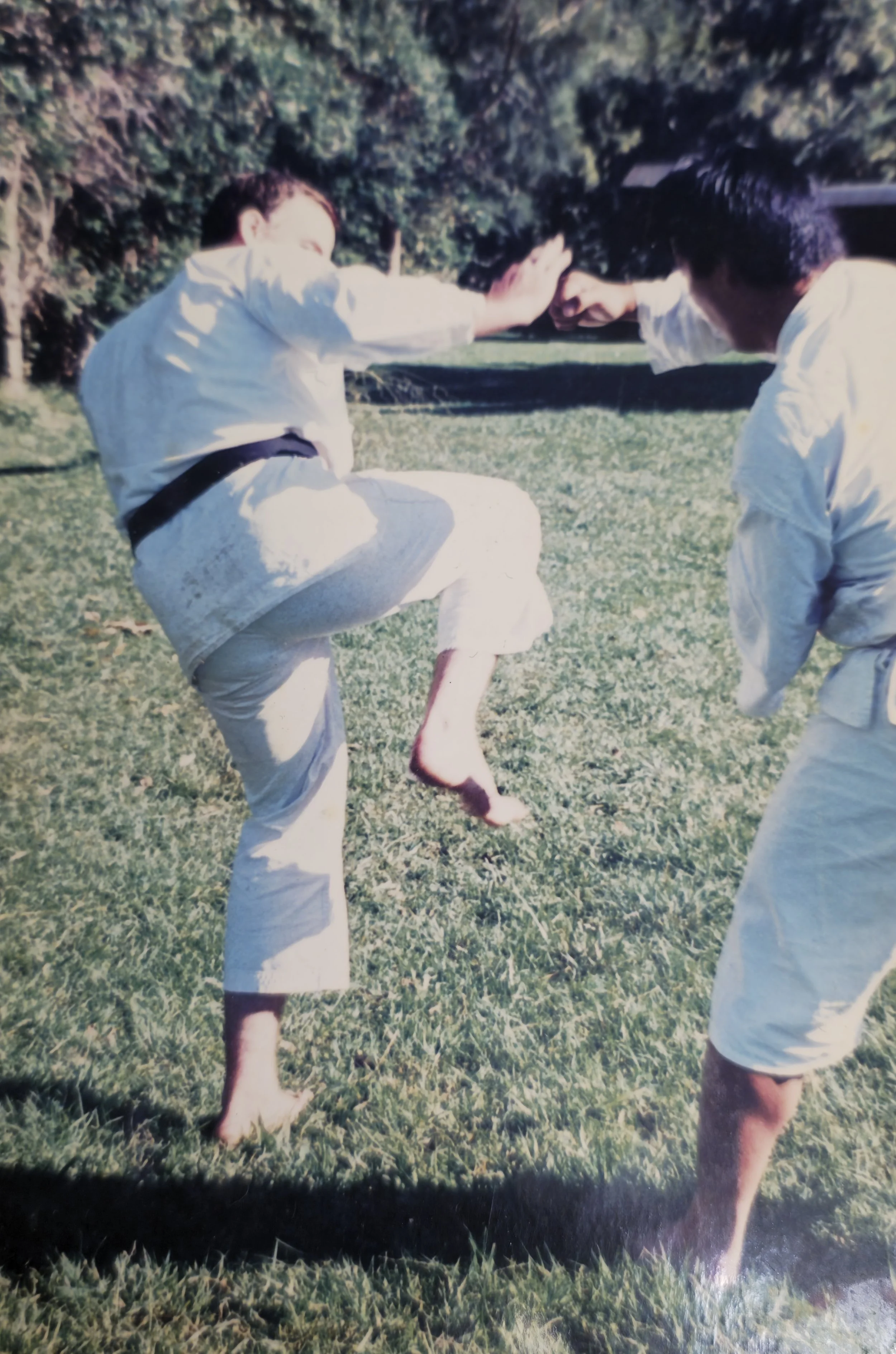Two martial artists practicing a technique outdoors on grass, wearing white uniforms, with one raising a leg and the other blocking with an arm.