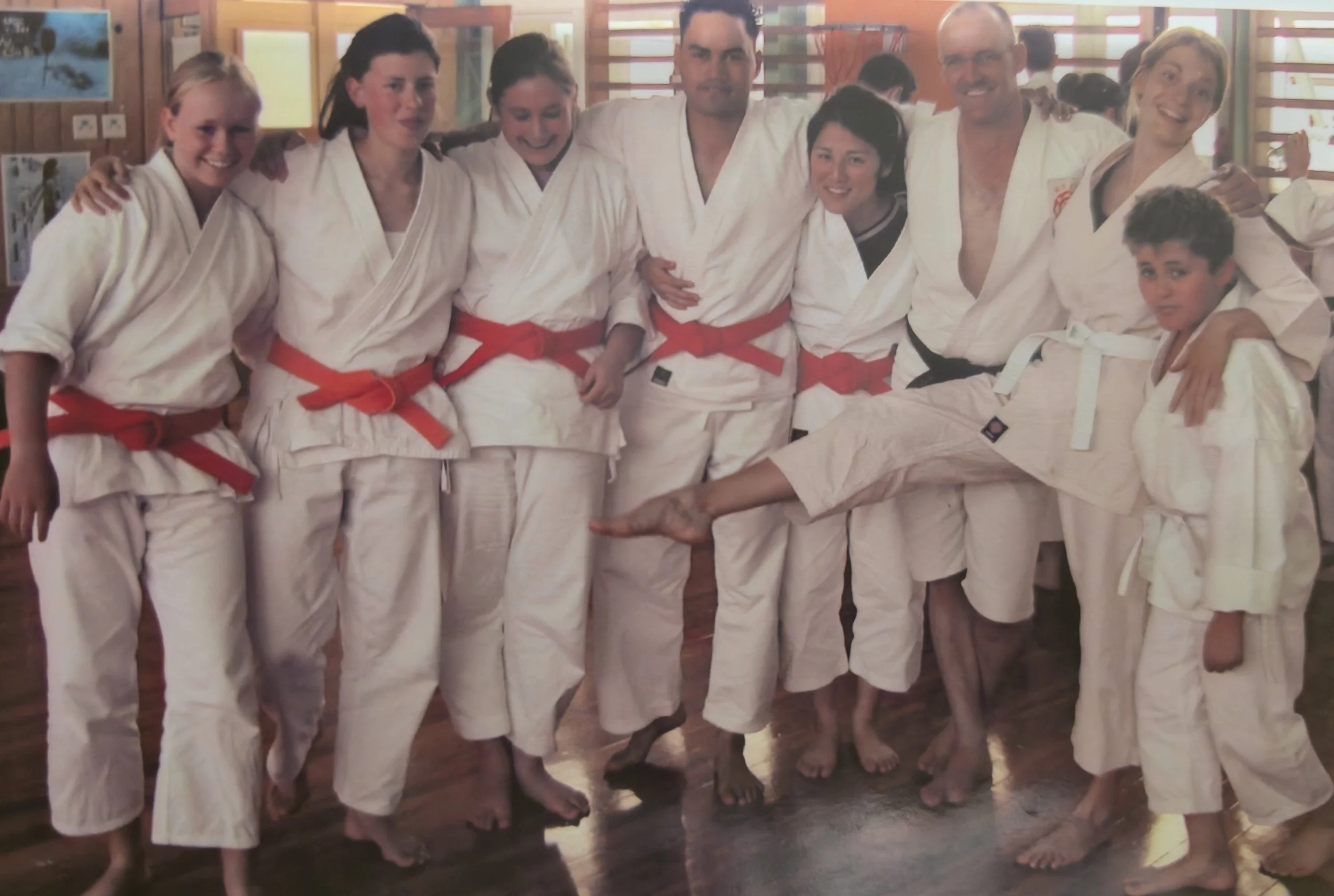 A group of people dressed in karate gis with red belts, standing together in a dojo, some smiling and one person doing a karate kick.