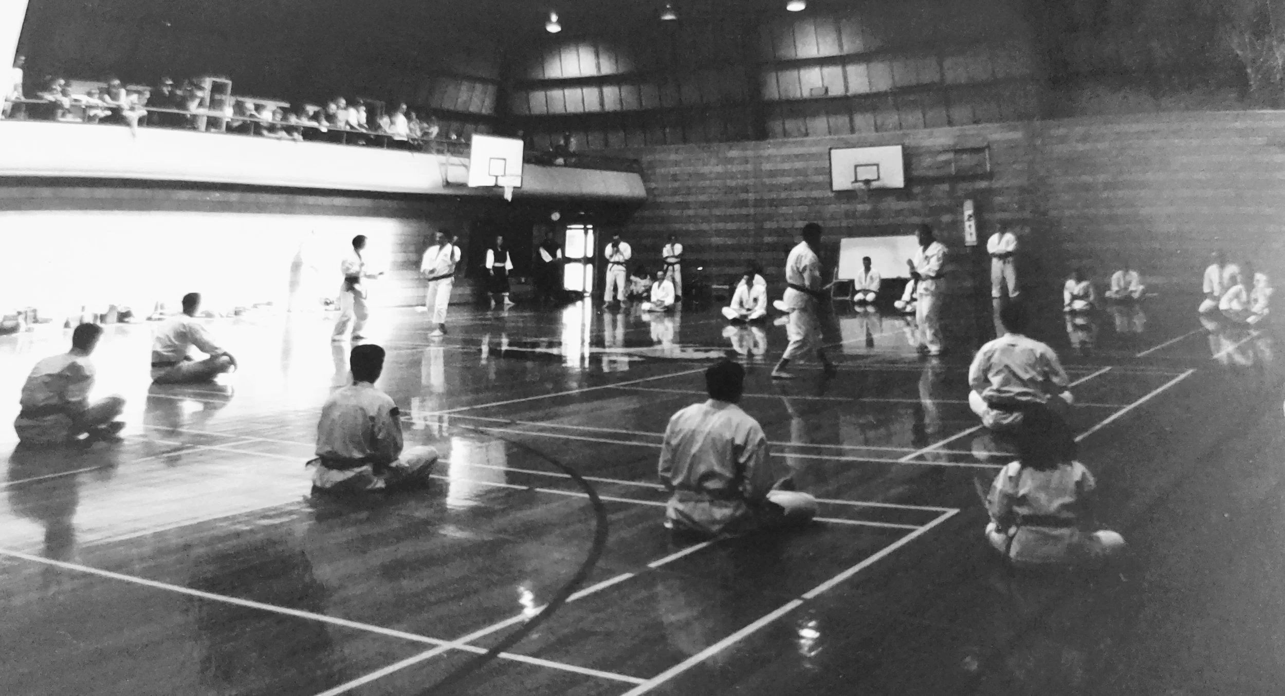 Black and white photo of a karate class in a gymnasium with students sitting in seiza position on the floor and instructors demonstrating techniques.