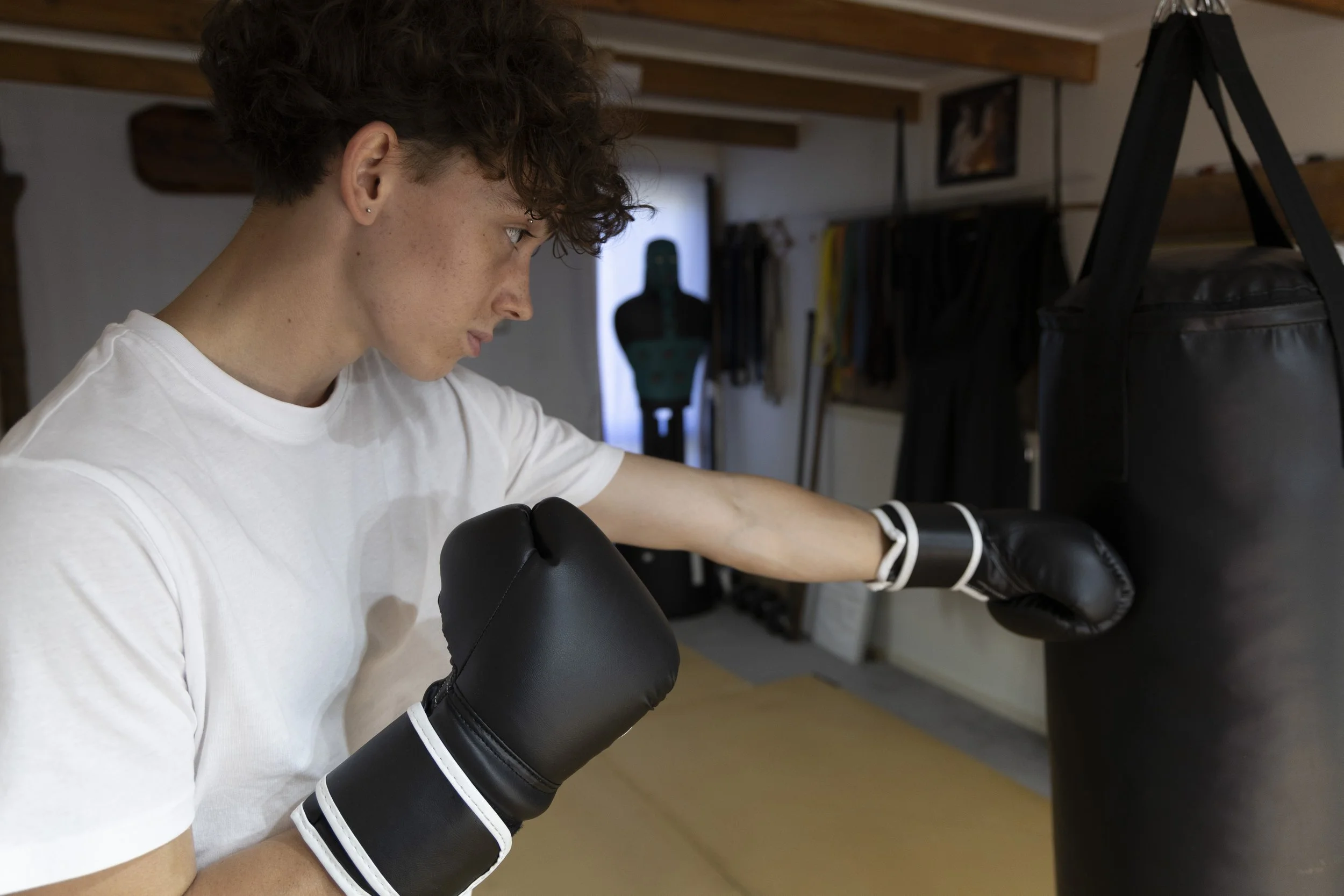 Young man practicing boxing with punching bag in a gym, wearing black boxing gloves and white T-shirt, focused on punching the bag.