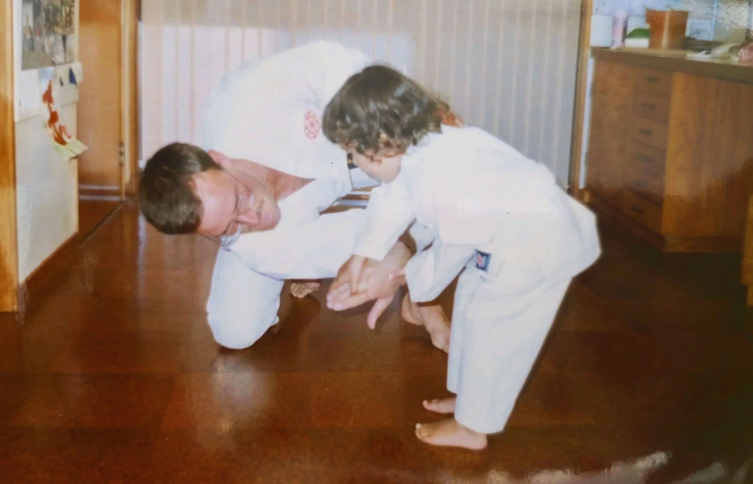 A man and a young boy practicing martial arts on a wooden floor in a room with wooden furniture.