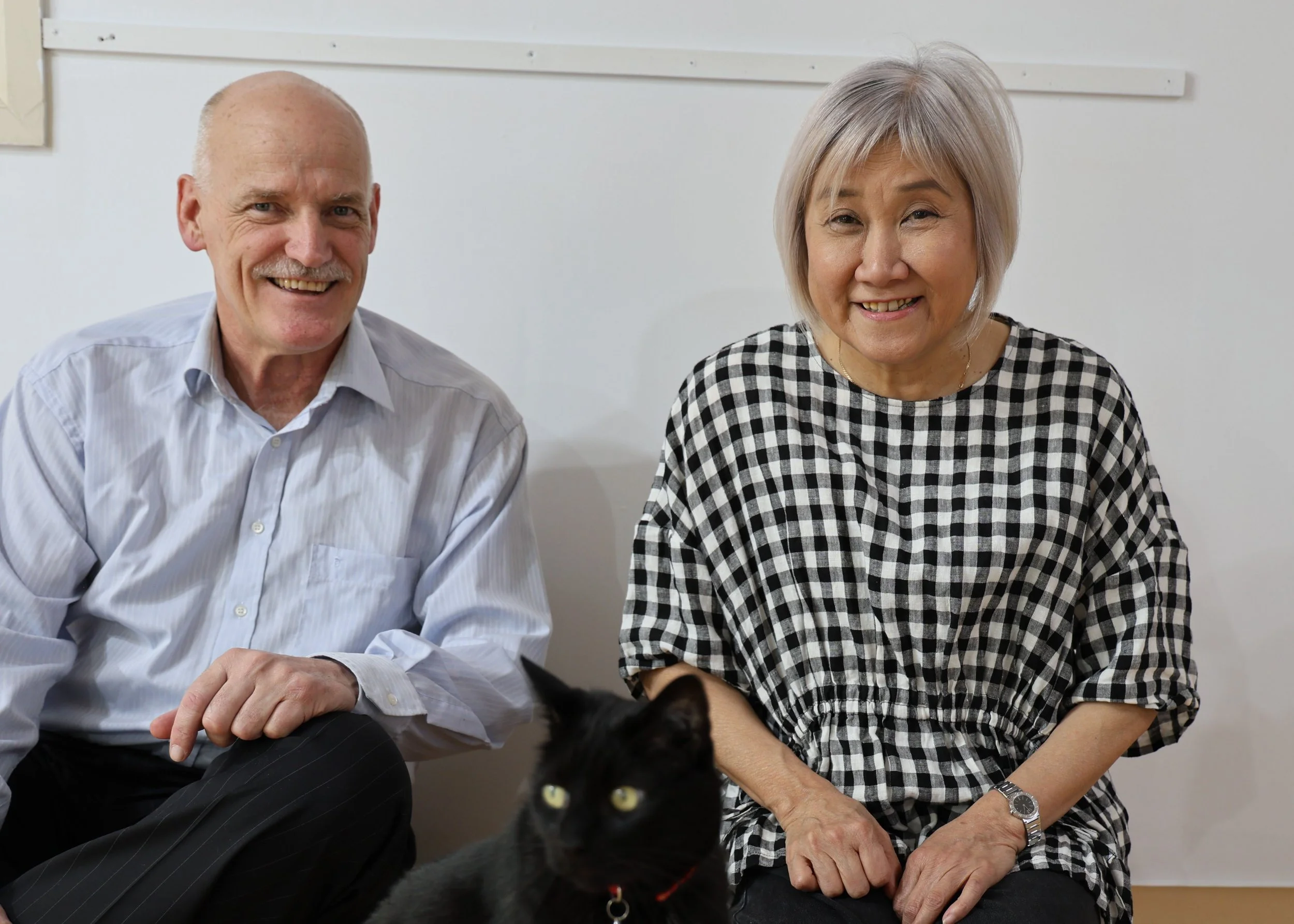 A smiling elderly man and woman sitting on the floor with a black cat in front of them, against a plain white wall.