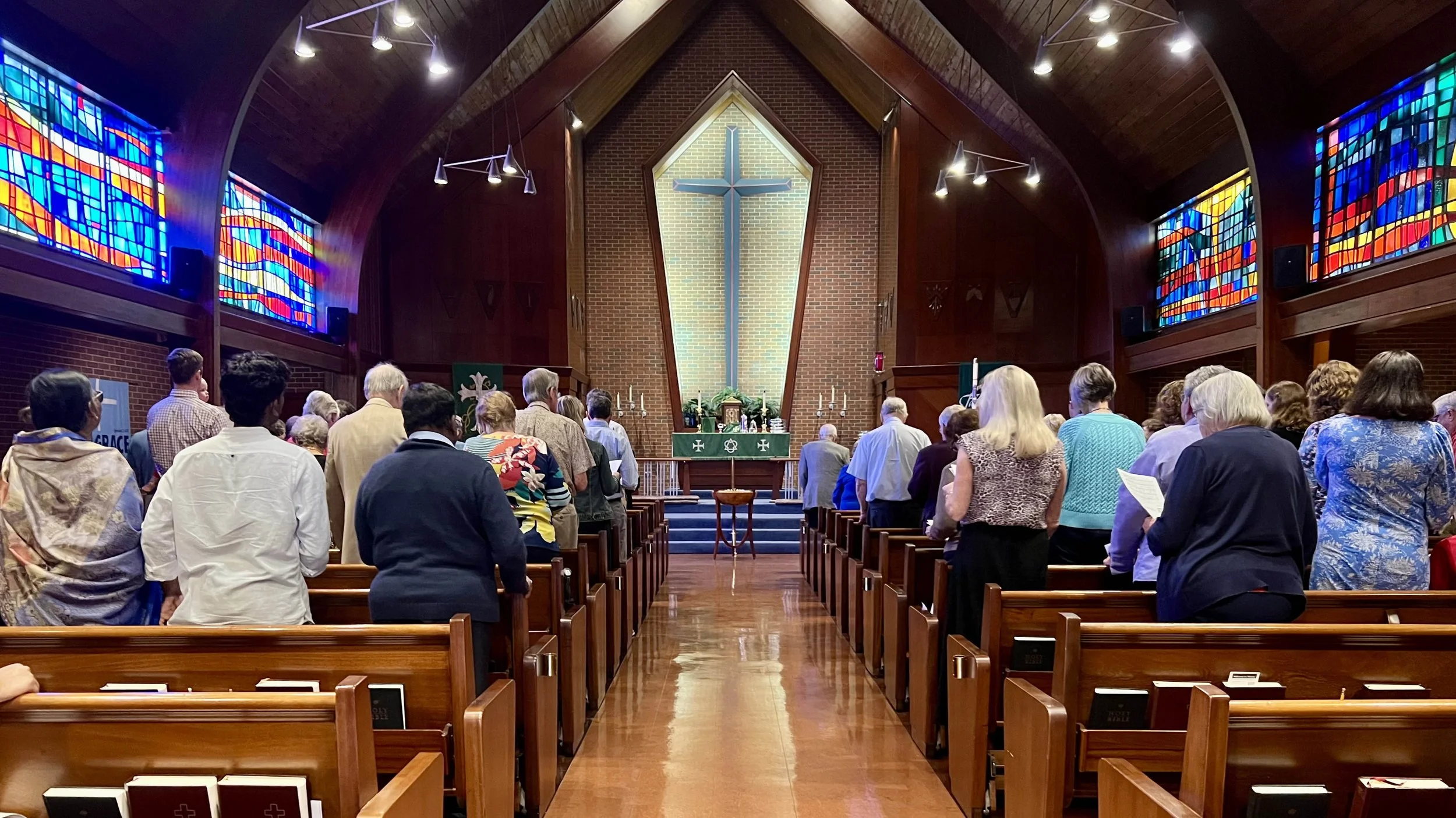 People gathered in a church for a service, standing in wooden pews facing the altar with a large cross behind it, stained glass windows on the sides, and dim lighting.