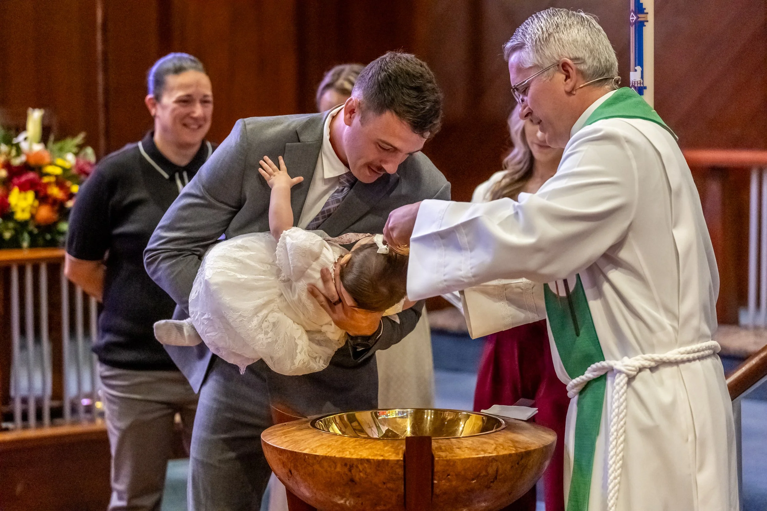 A baptism ceremony taking place in a church, with a priest pouring water over a baby held by a man, while another woman looks on, and a woman in the background smiling.