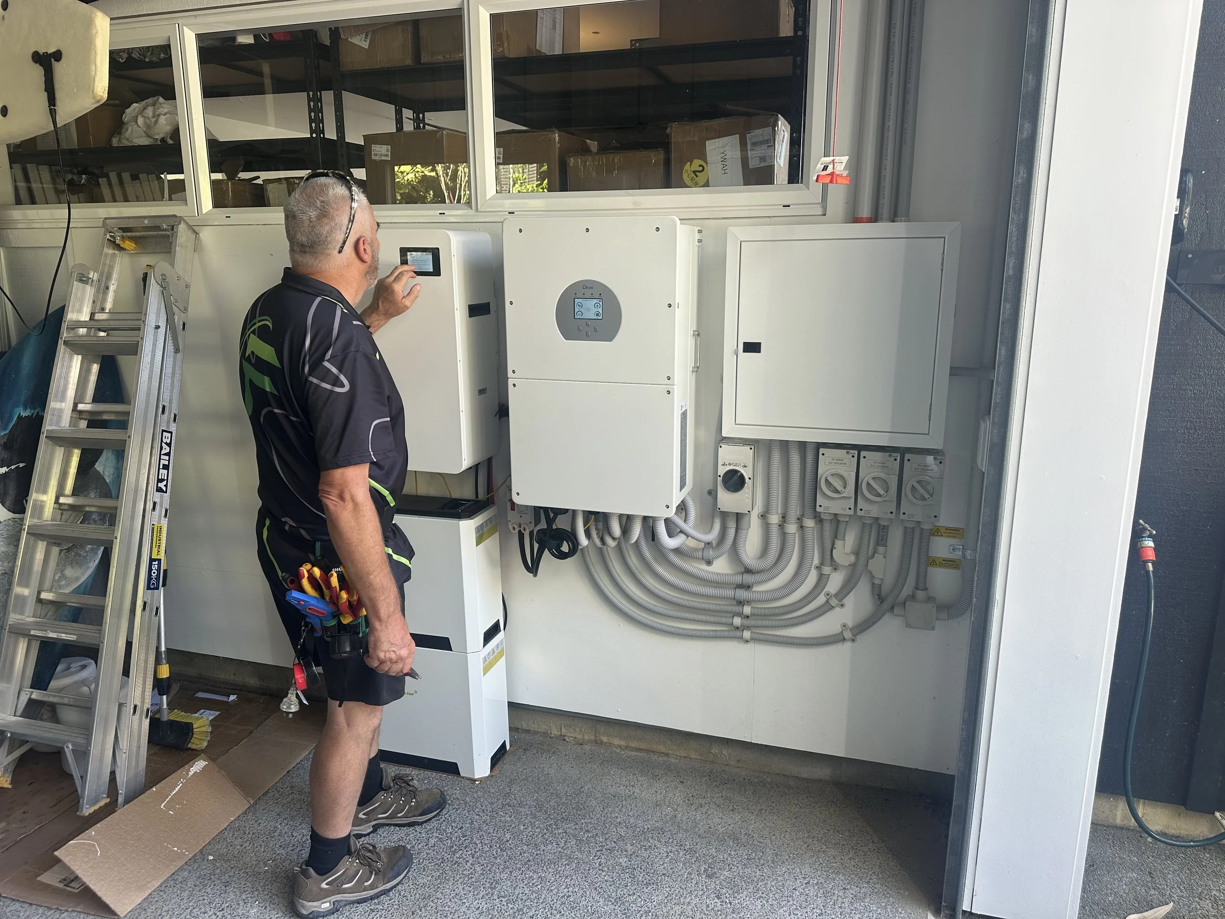 A worker in black work attire with tools attached to his waist is interacting with an electronic control panel by a large white industrial machine outside of a building. There is a ladder leaning against the wall nearby.