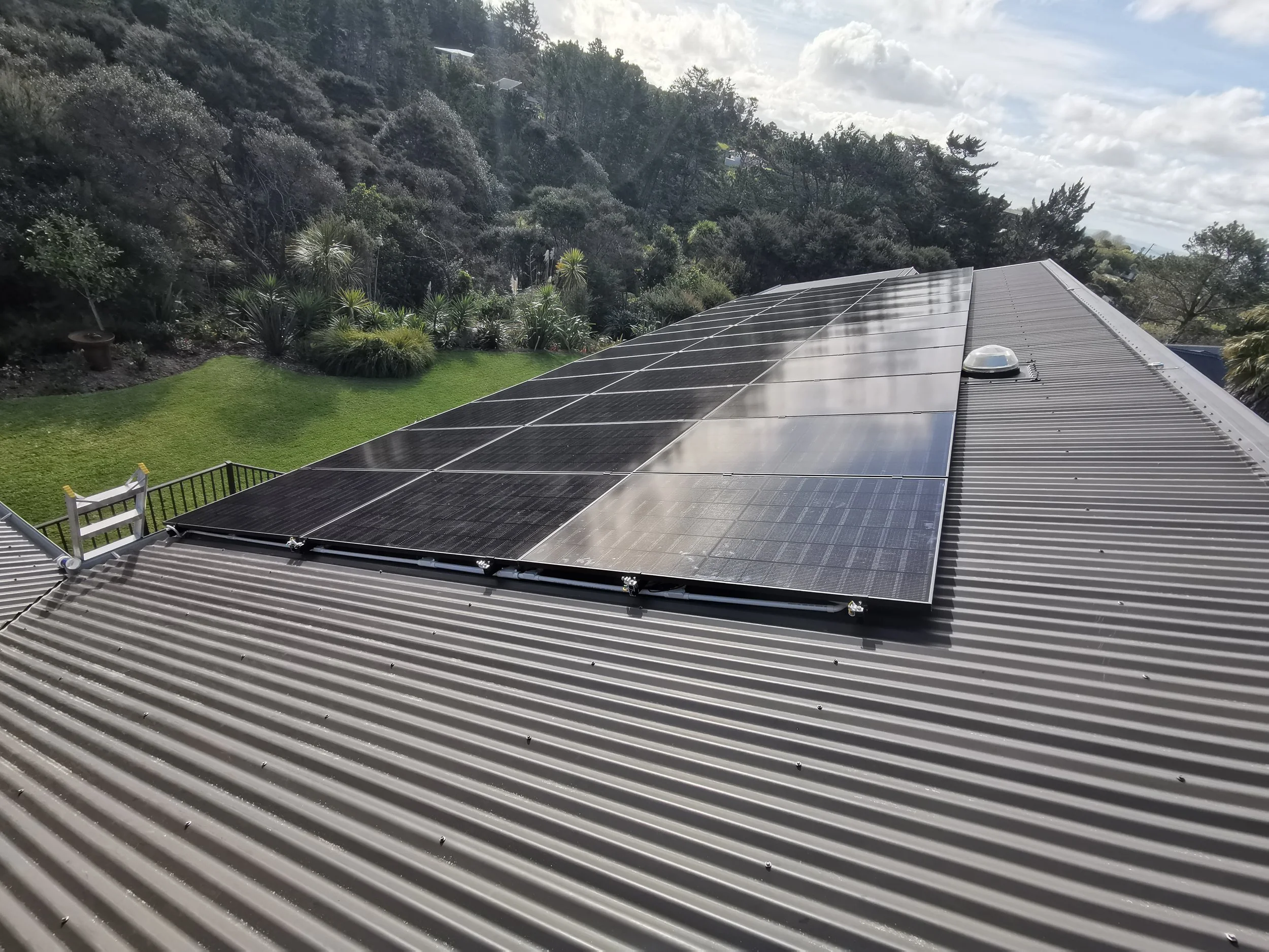 Solar panels installed on corrugated metal roof in a backyard with green lawn and trees in the background.