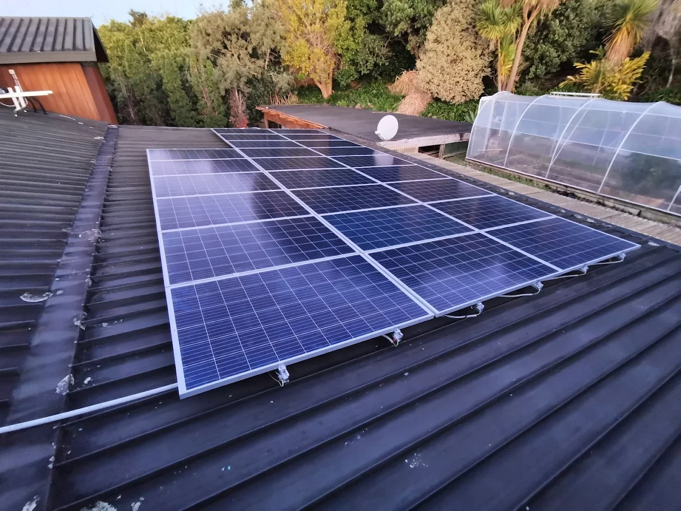 Solar panels installed on a sloped black metal roof with a garden and trees in the background.