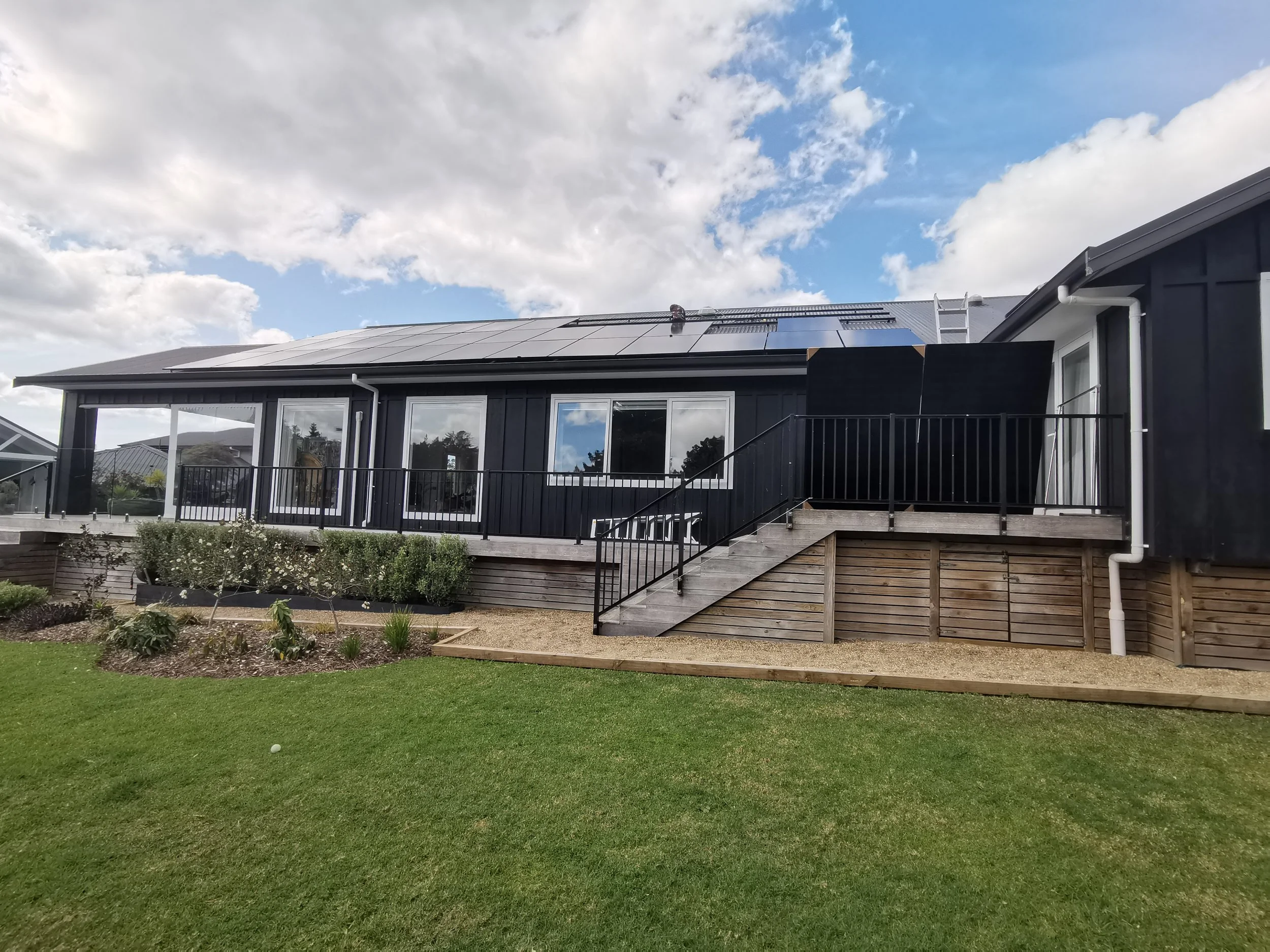 A black house with a solar panel roof, steps leading up to a deck, and a landscaped yard with grass and plants.