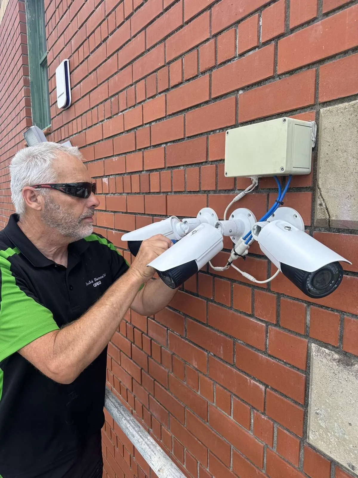 A technician wearing sunglasses and a black and green uniform working on outdoor security cameras mounted on a brick wall.