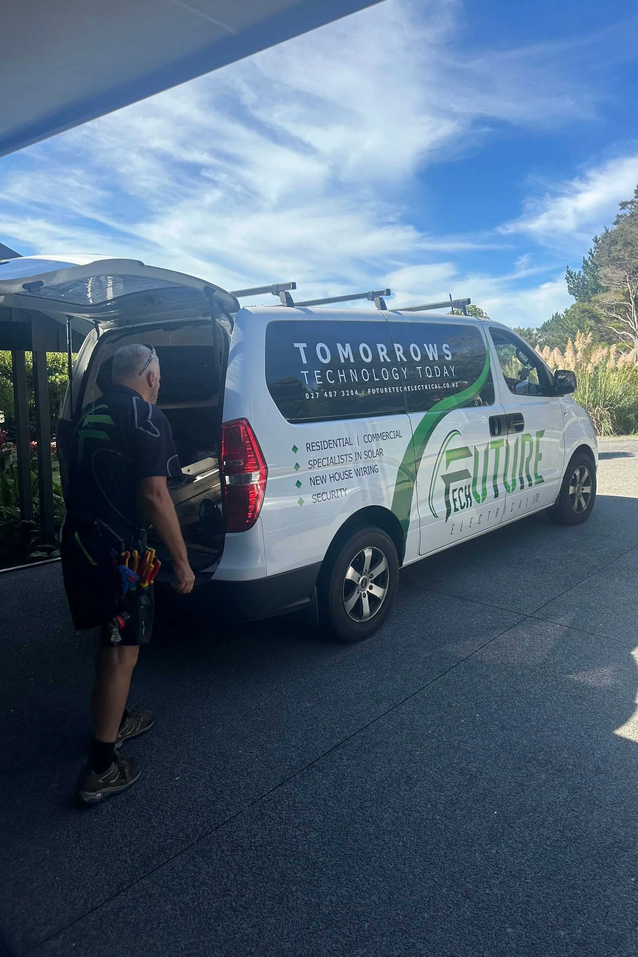 A technician unloading equipment from a white company van with green and black lettering, parked outdoors on a sunny day. The van has the words 'Future Electrical' and services listed, including residential, commercial, solar, house wiring, and security.