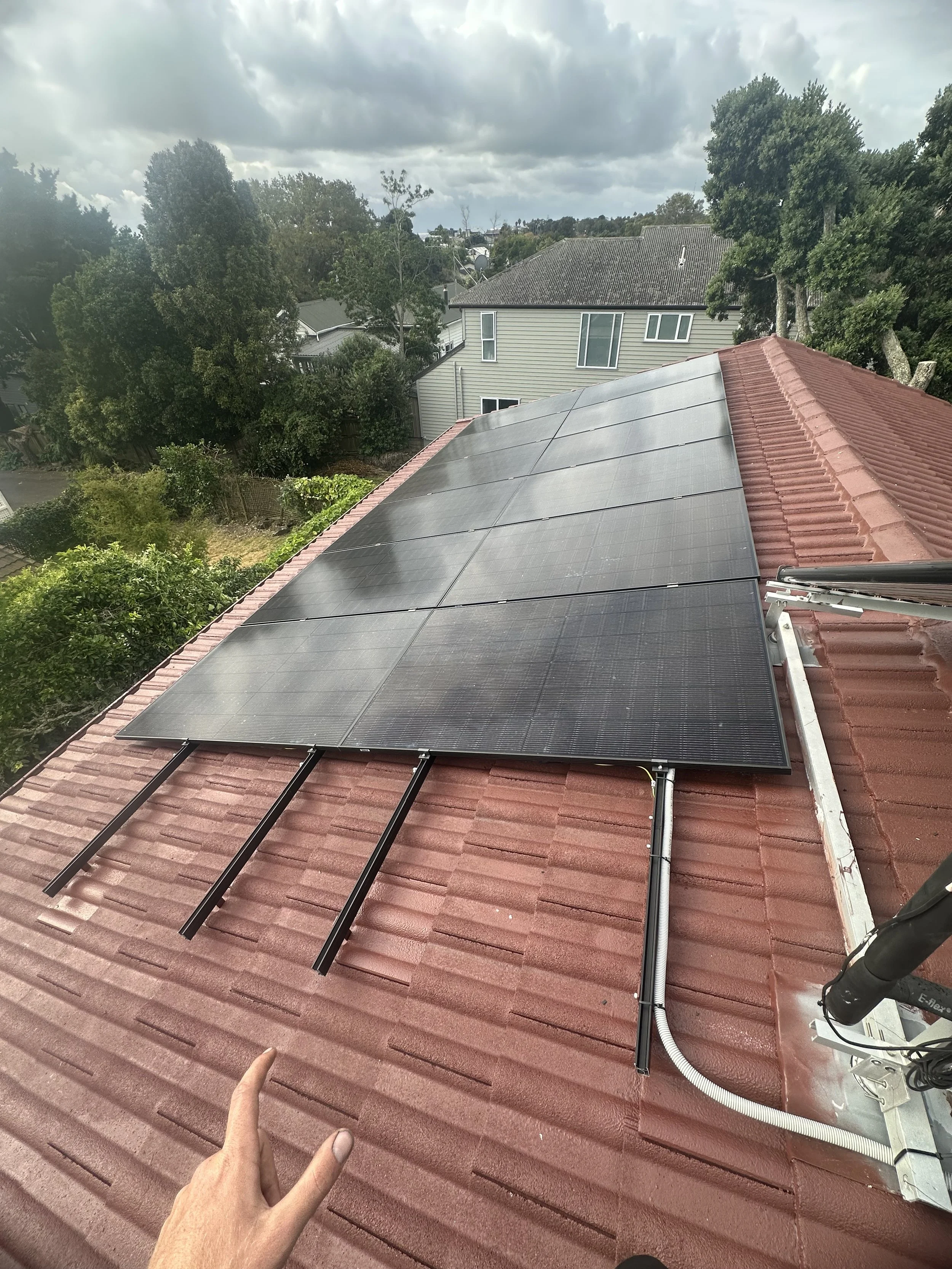 Photograph of solar panels installed on a red-tiled roof of a house, with a person's hand visible in the bottom left corner pointing towards the panels.