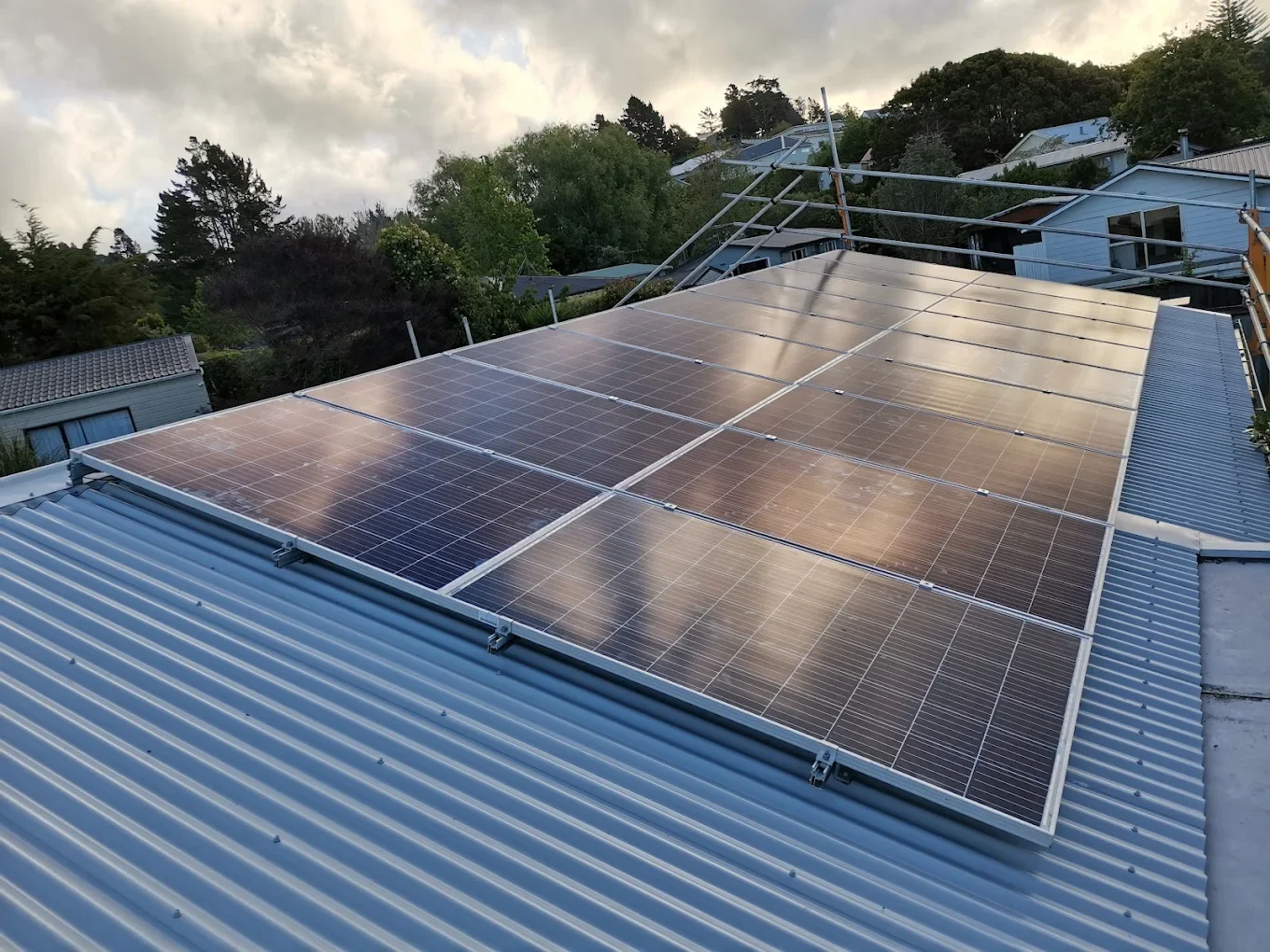 Solar panels installed on a corrugated metal roof, with a backdrop of trees and houses under a cloudy sky.