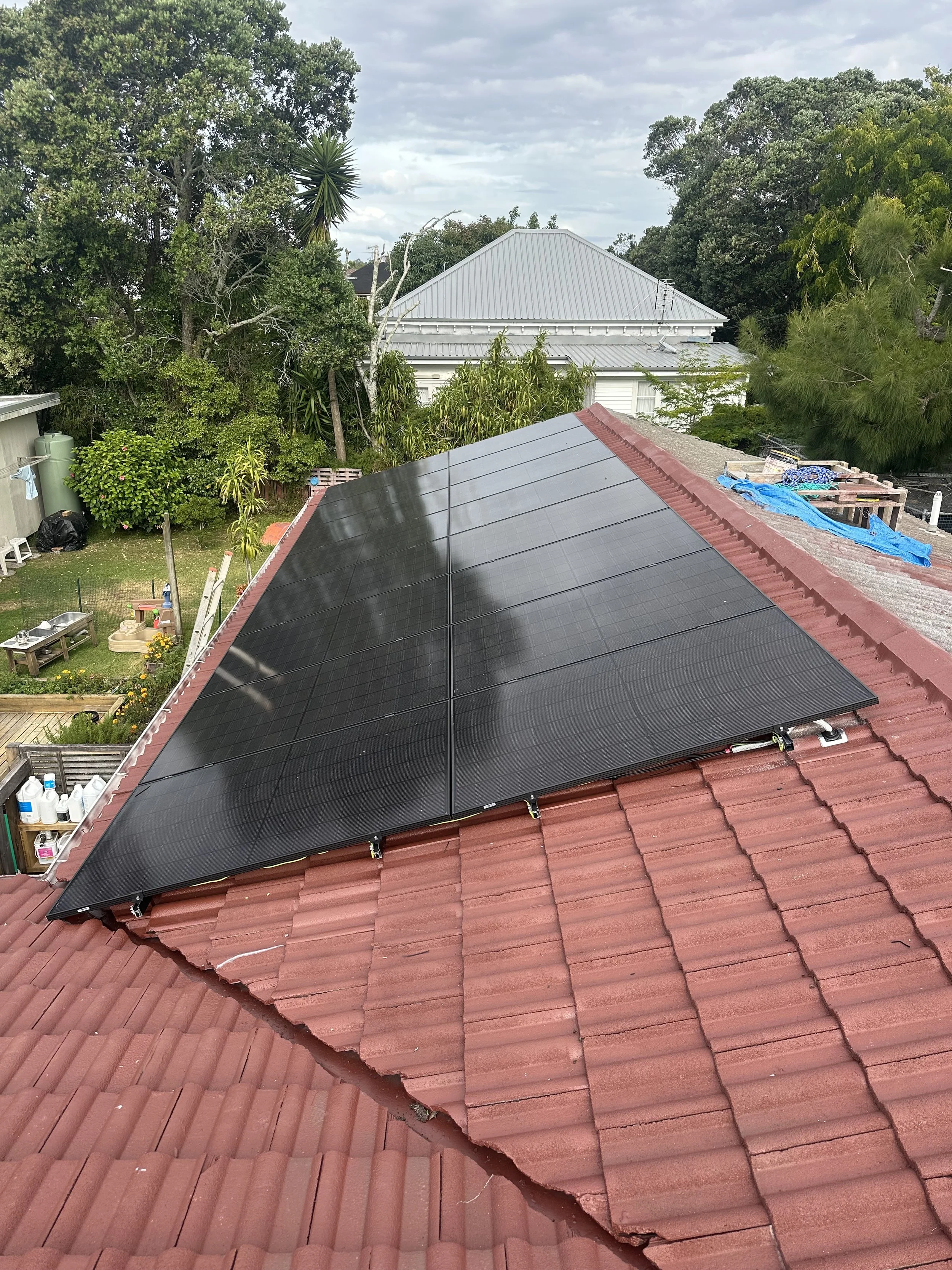 Solar panels installed on a red tile roof in a residential neighborhood with trees and cloudy sky in the background.