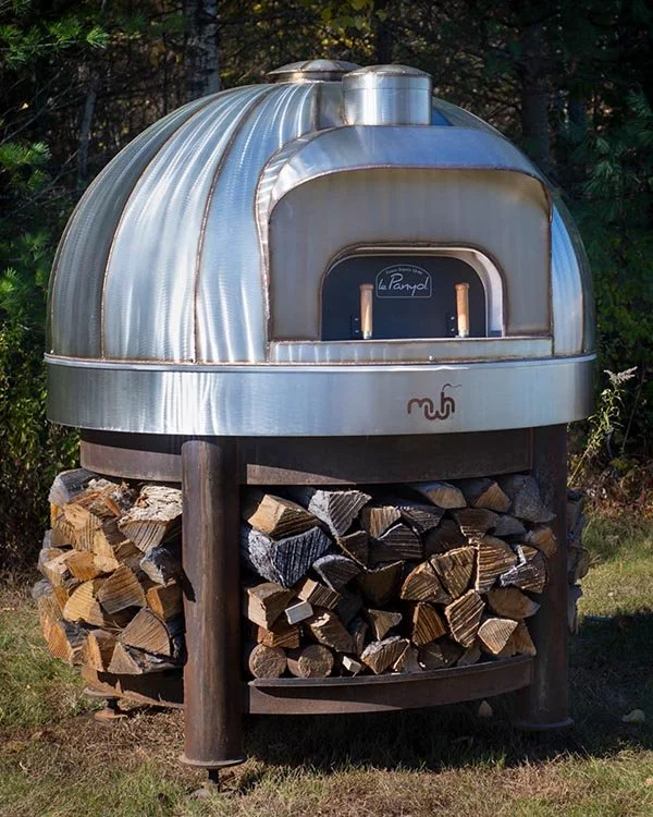 A wood-fired pizza oven with a metallic dome top, sitting on a wooden stand filled with cut firewood.