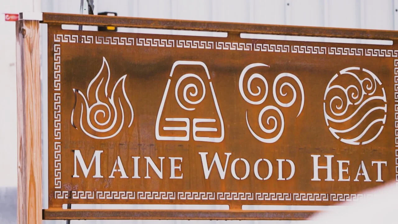 A metal sign for Maine Wood Heat with symbols representing fire, heat, wind, and air, framed by a decorative border. Wood-Fired Ovens.