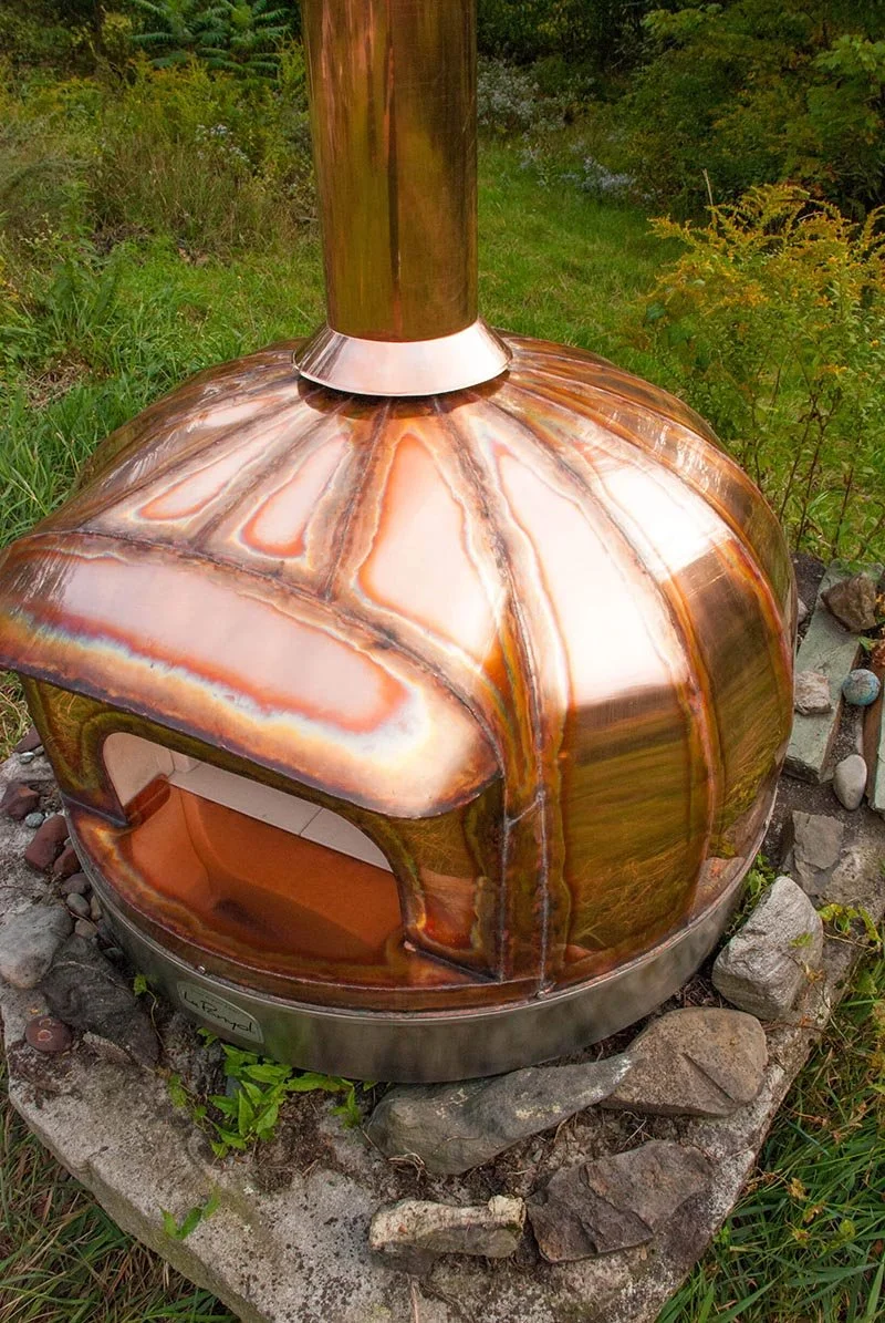 Backyard Wood Burning Oven with a center chimney, situated on a stone platform surrounded by green grass and plants.