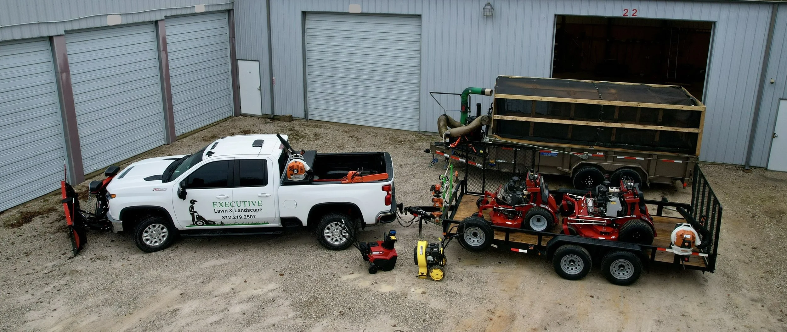 White pickup truck with lawn and landscape equipment attached, parked next to a trailer carrying more landscaping machinery, in front of a large gray building with closed garage doors.