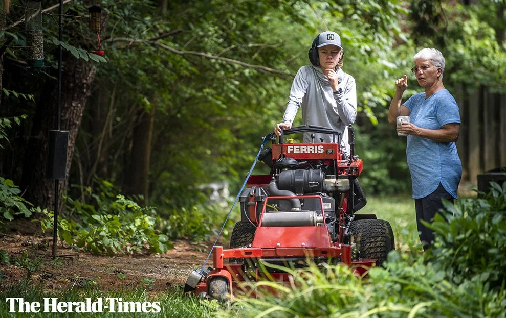 An elderly woman and a young man are standing outdoors in a wooded area near a red lawn mower, having a conversation.