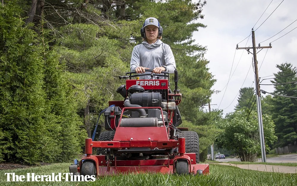 A young person mowing the grass with a red Ferris riding lawn mower, wearing a gray hoodie, helmet, and headphones, with green bushes and trees in the background.