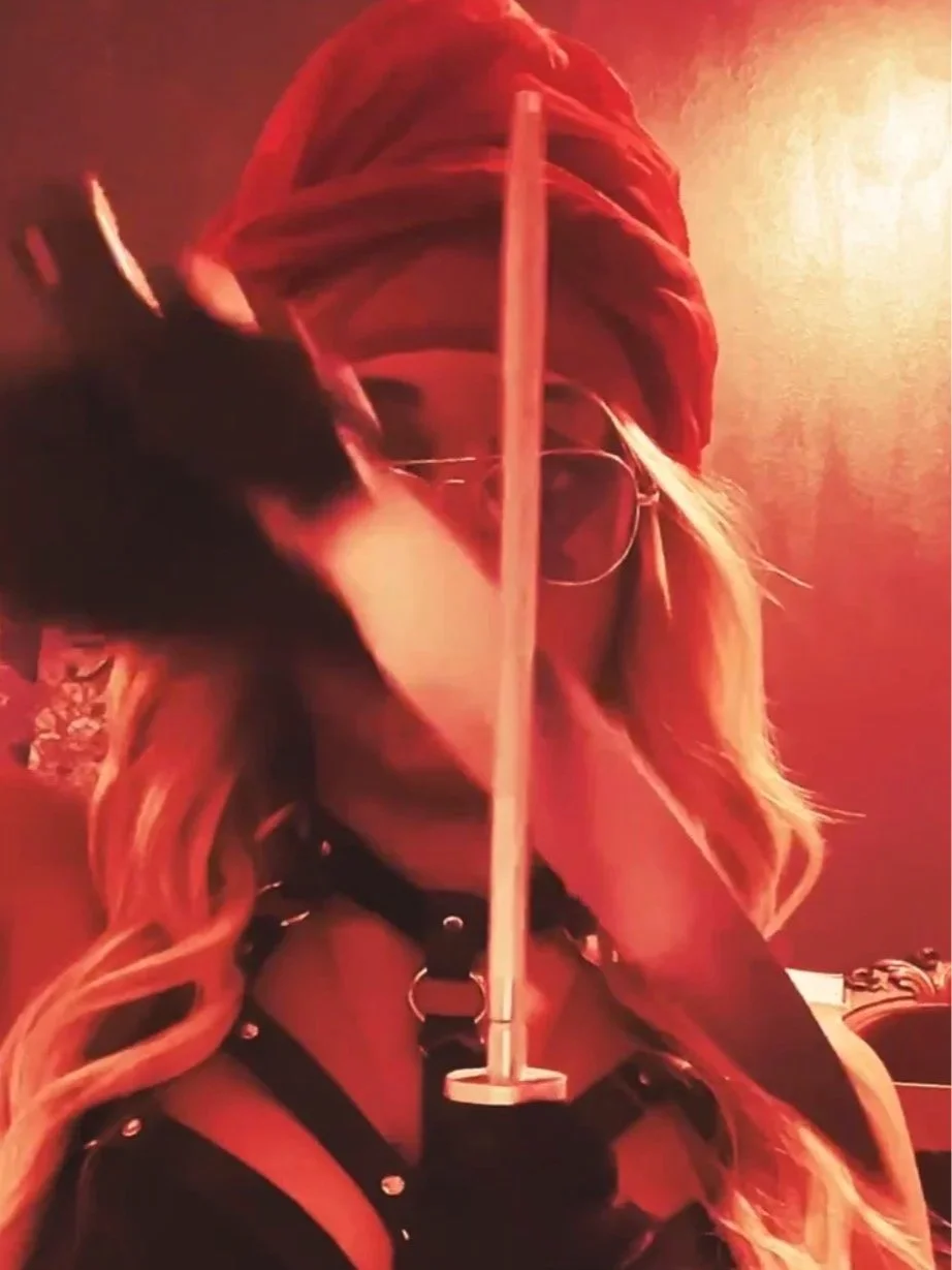 A woman with long blonde hair wearing glasses, a red headscarf, leather harness, and gloves, holding a sword vertically in front of her face in a dim, reddish lighting environment.