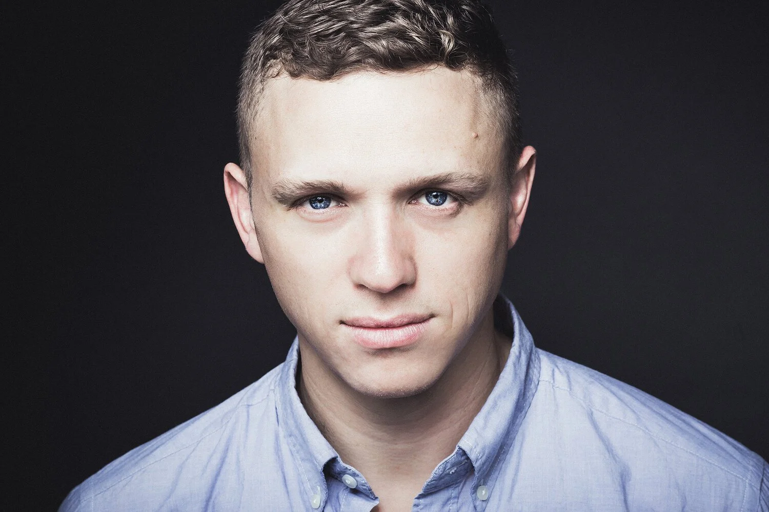 Portrait of Owner, Producer, and Director Tobin Del Cuore - a man with short hair and blue eyes, wearing a light blue shirt on a black background.