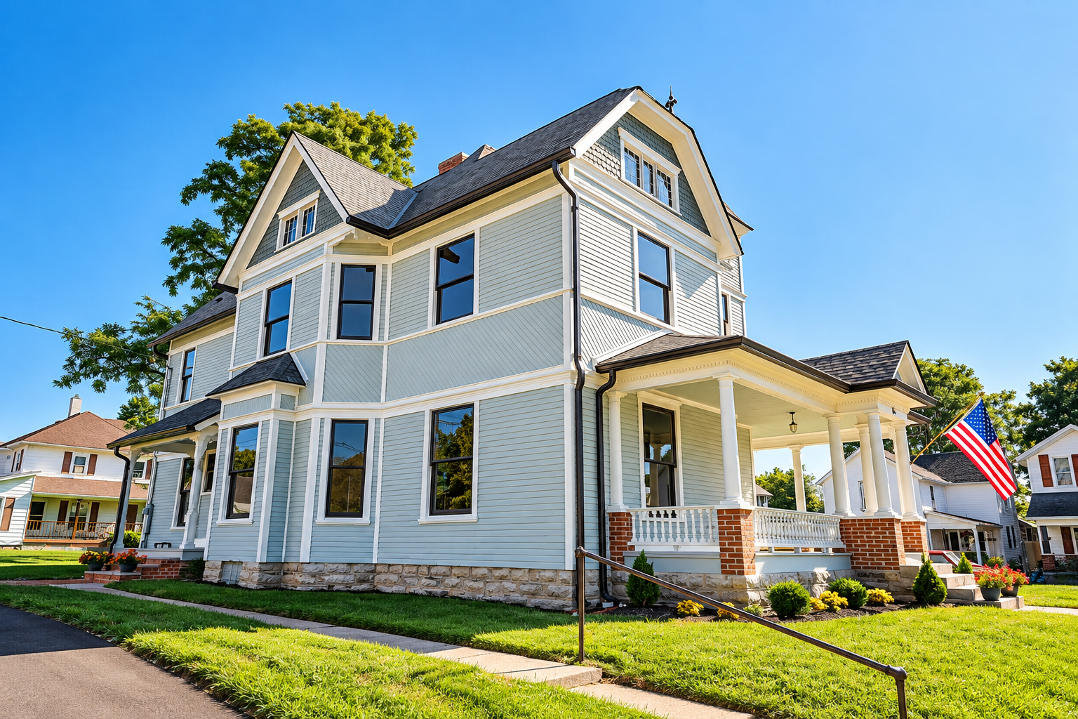 Large historic American home with light blue siding, white trim, wraparound porch, landscaped lawn, and American flag under a clear blue sky