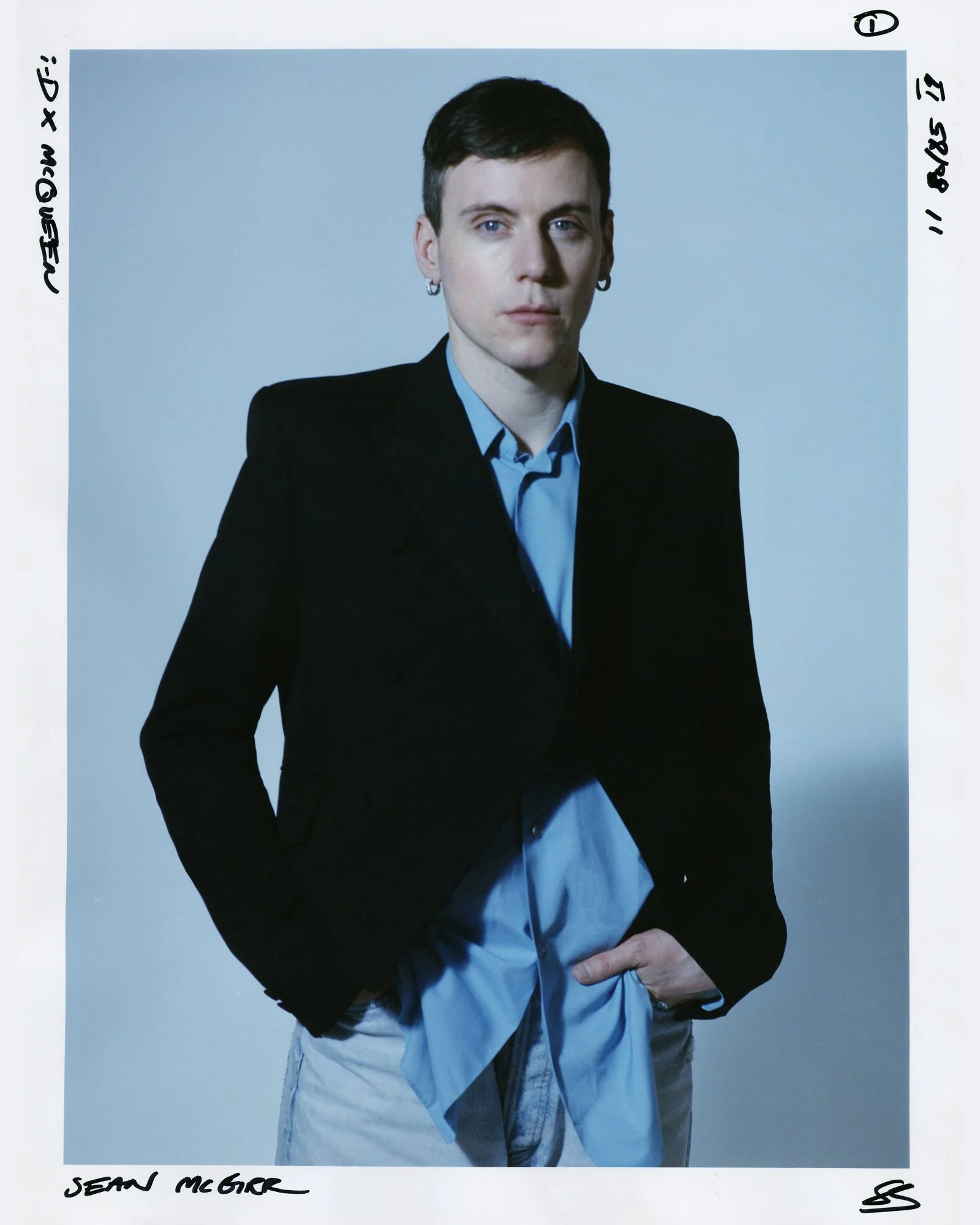 Portrait of a young man with short dark hair, wearing a black blazer over a light blue shirt. He has earrings and is posed with a serious expression, standing against a plain light background.