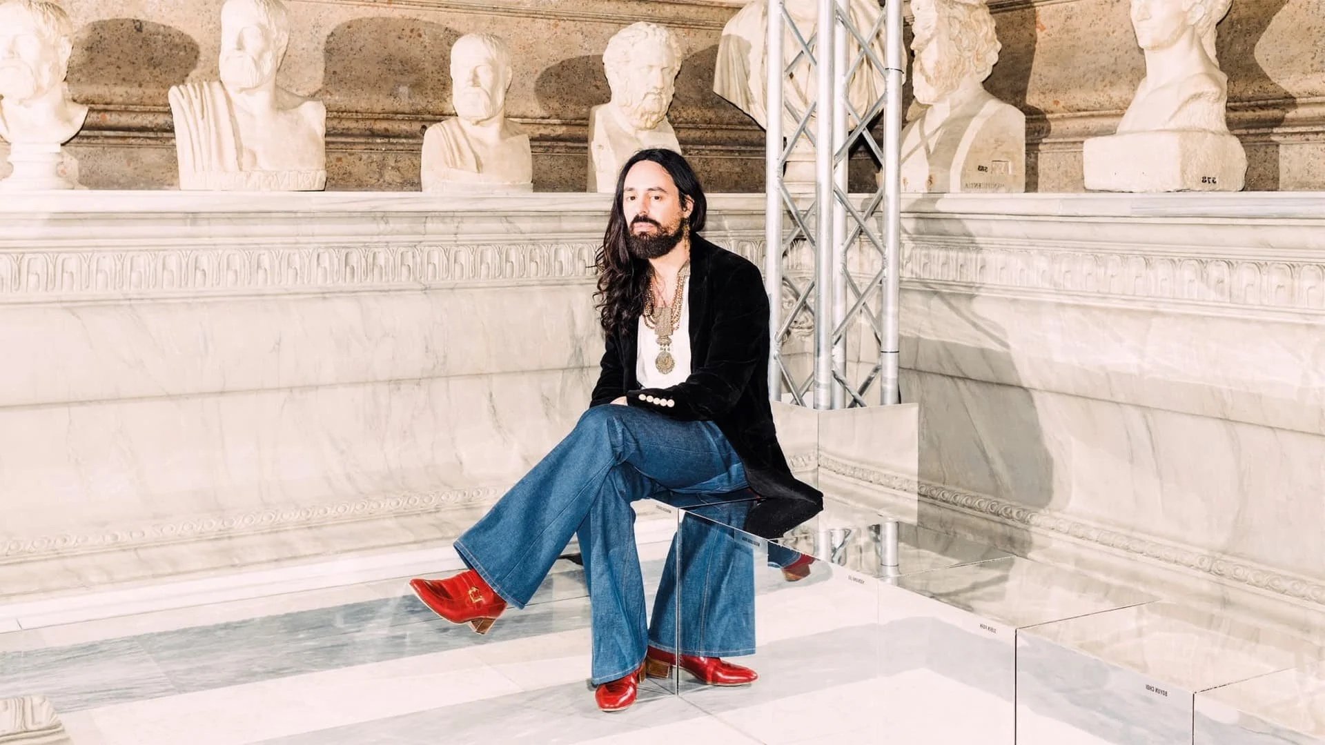 A man with long dark hair, a beard, and light skin, dressed in a black blazer, white shirt, wide-leg blue jeans, and red heeled boots, sitting on a modern glass bench in a museum gallery with classical bust sculptures on the wall behind him.